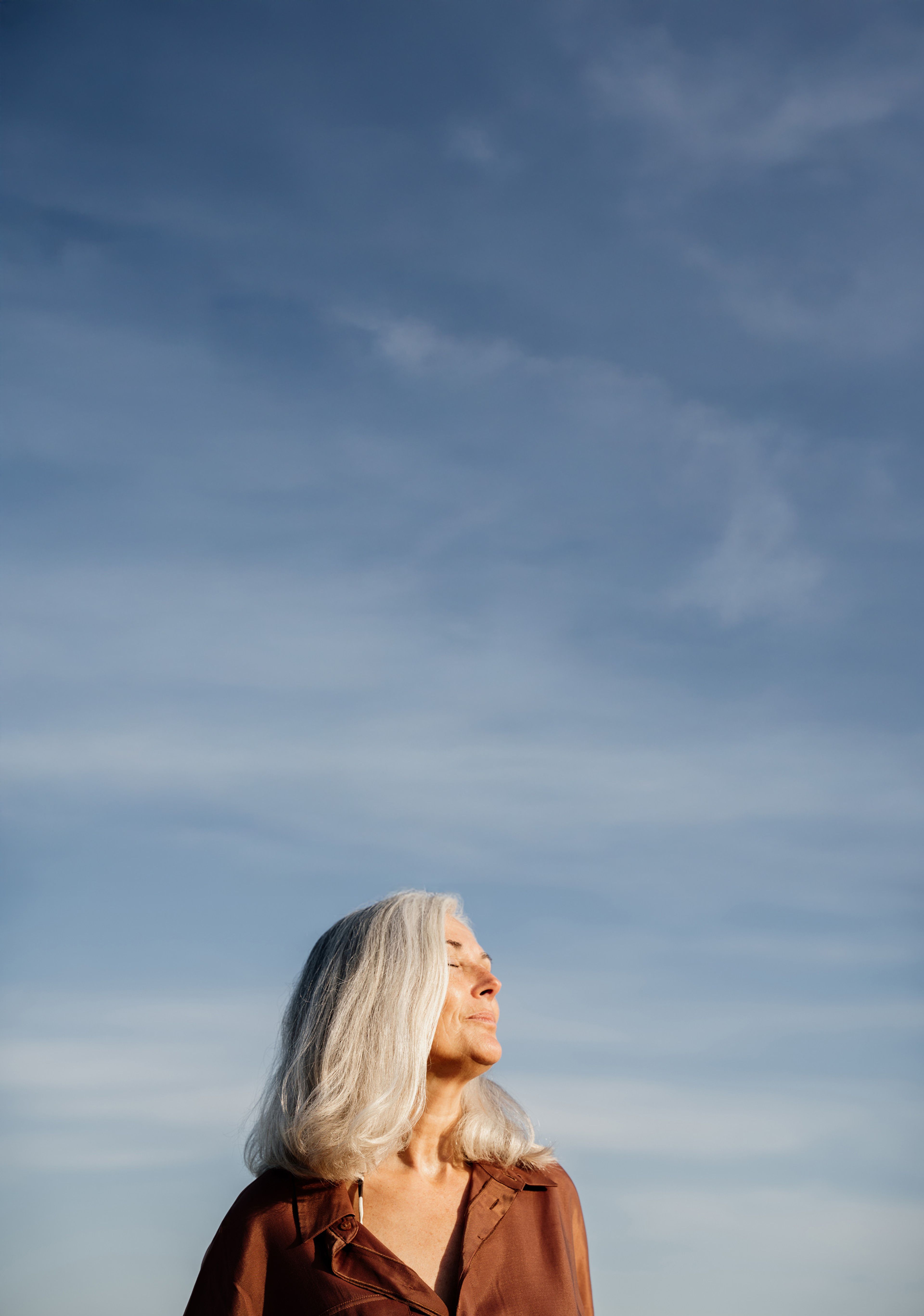A woman in a brown shirt, looking up and basking in the afternoon sun with her eyes closed, with blue skies all around
