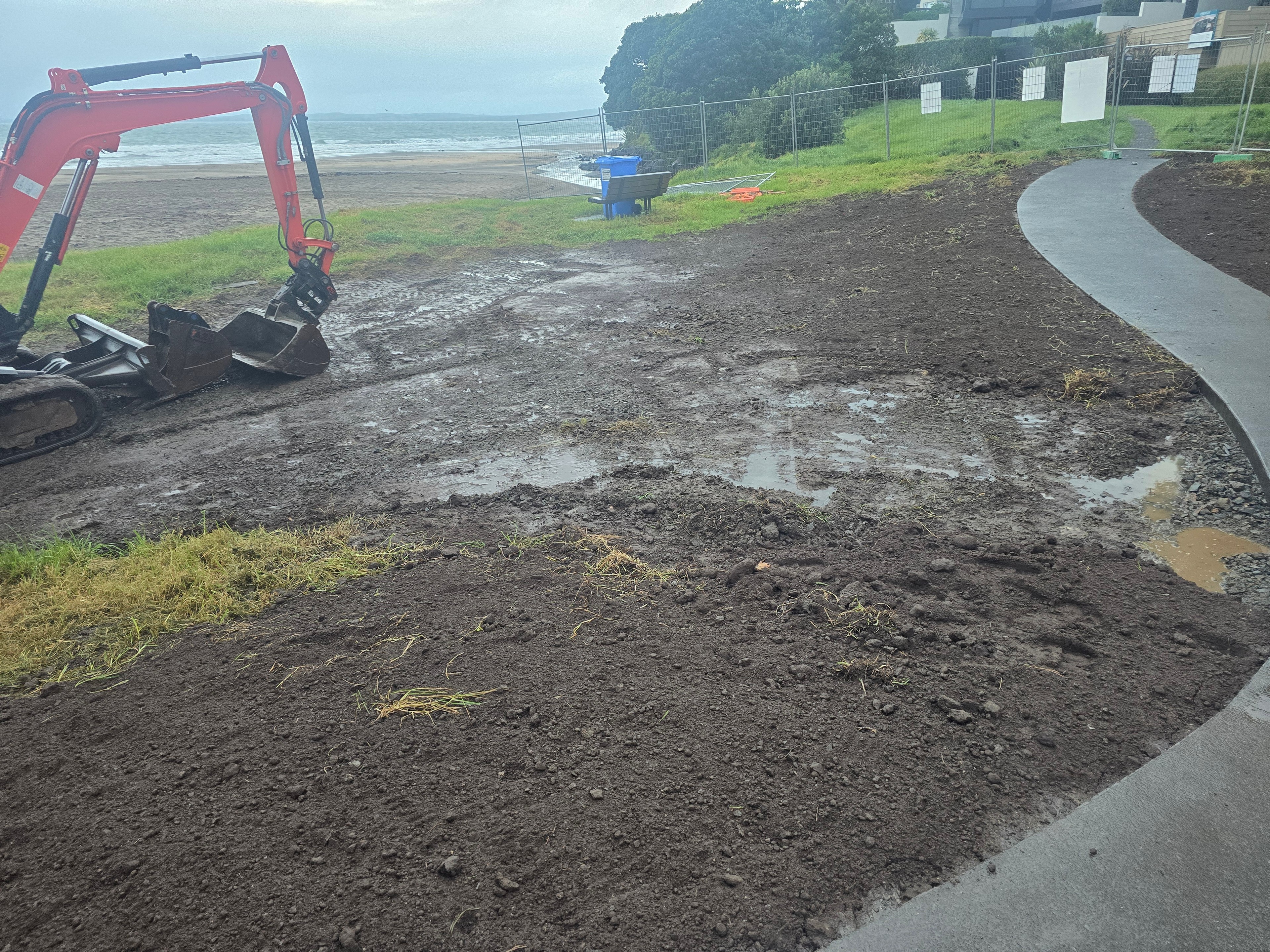 Excavator shaping topsoil alongside a new curved concrete path, with the beach behind
