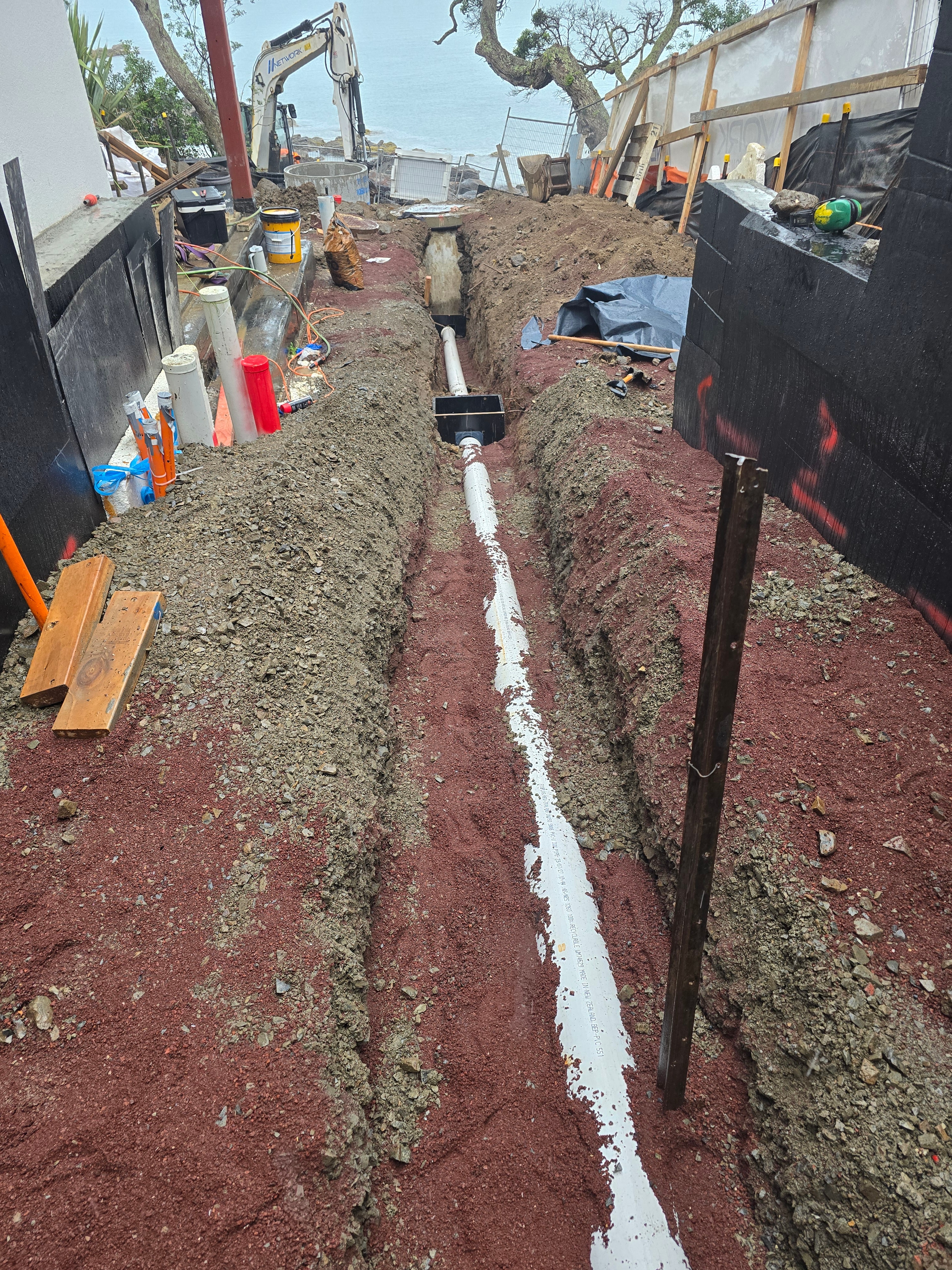 Stormwater trench with PVC pipe laid to fall on scoria bedding, running toward the excavator and the sea