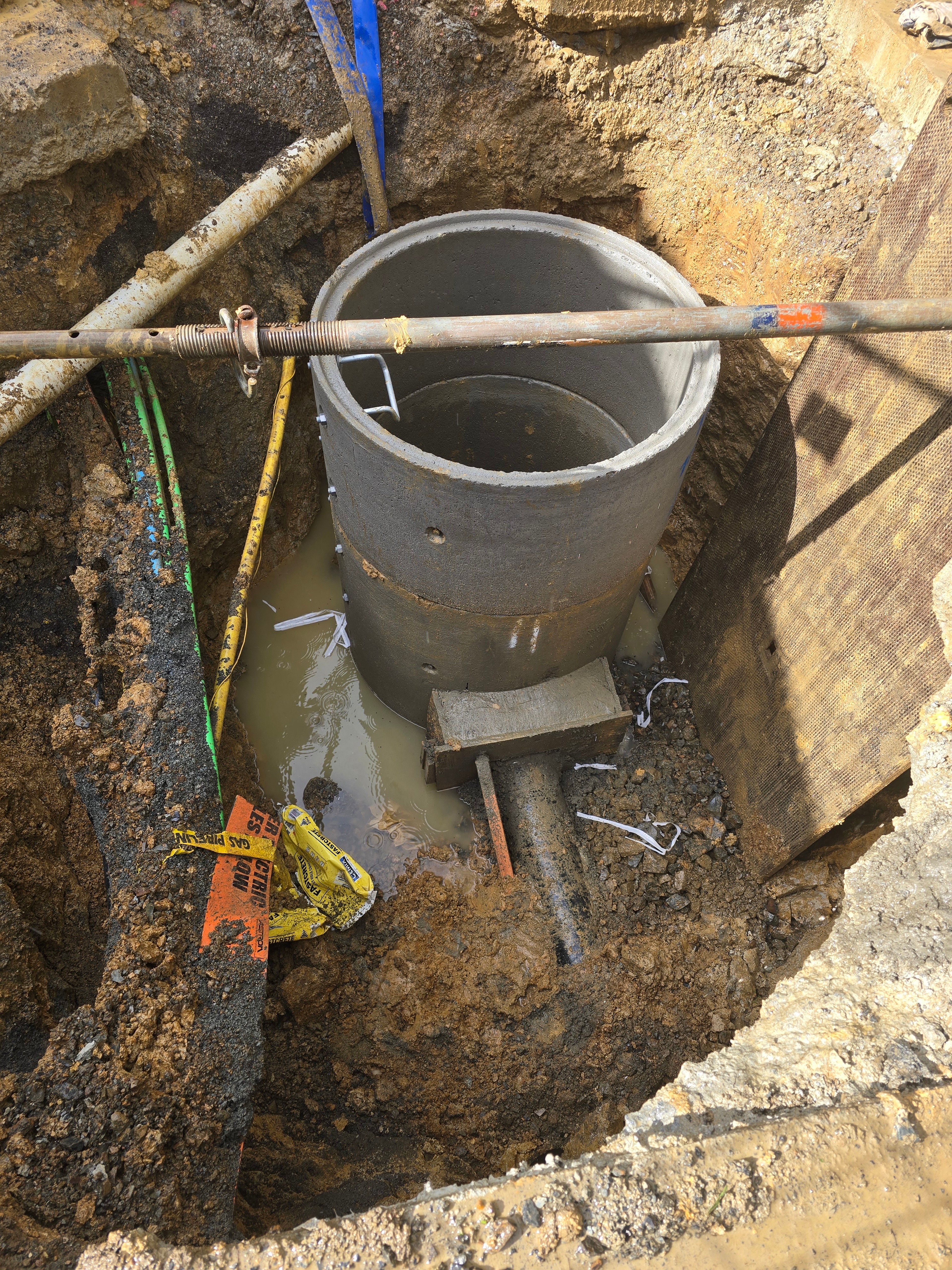 New concrete manhole chamber being set into an excavated pit, with the incoming pipe and buried service markers exposed