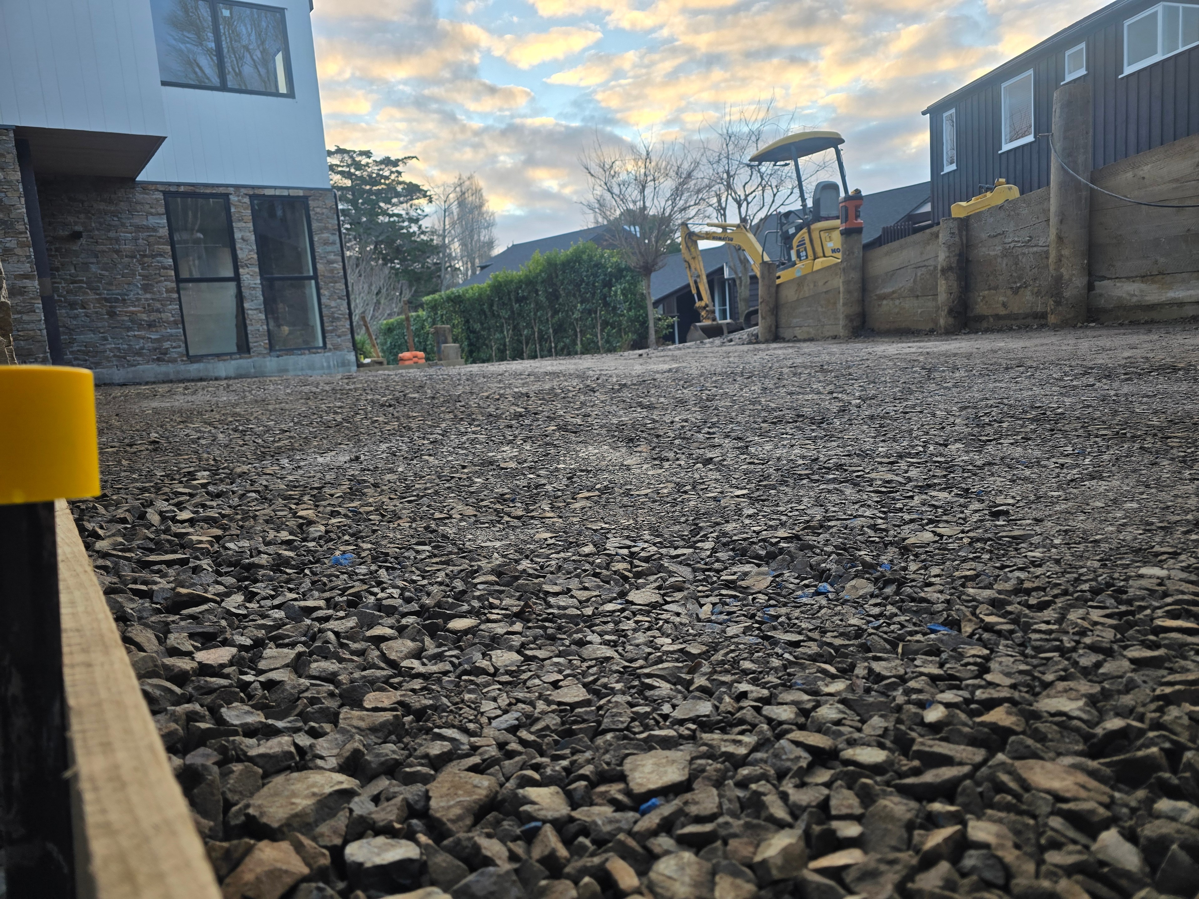 Excavator parked on a finished basecourse courtyard at dusk, timber retaining wall alongside