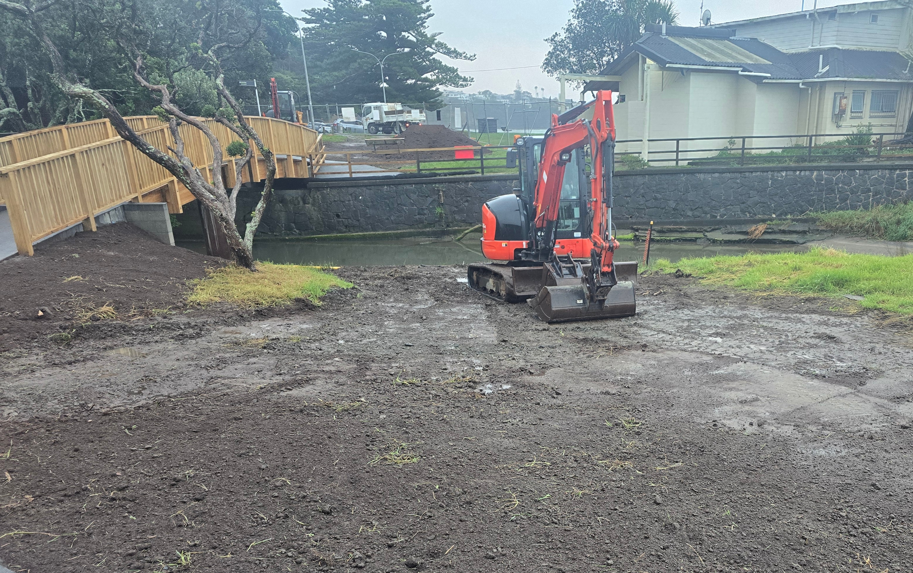 Excavator standing on freshly graded ground beside a completed footbridge and stone retaining wall