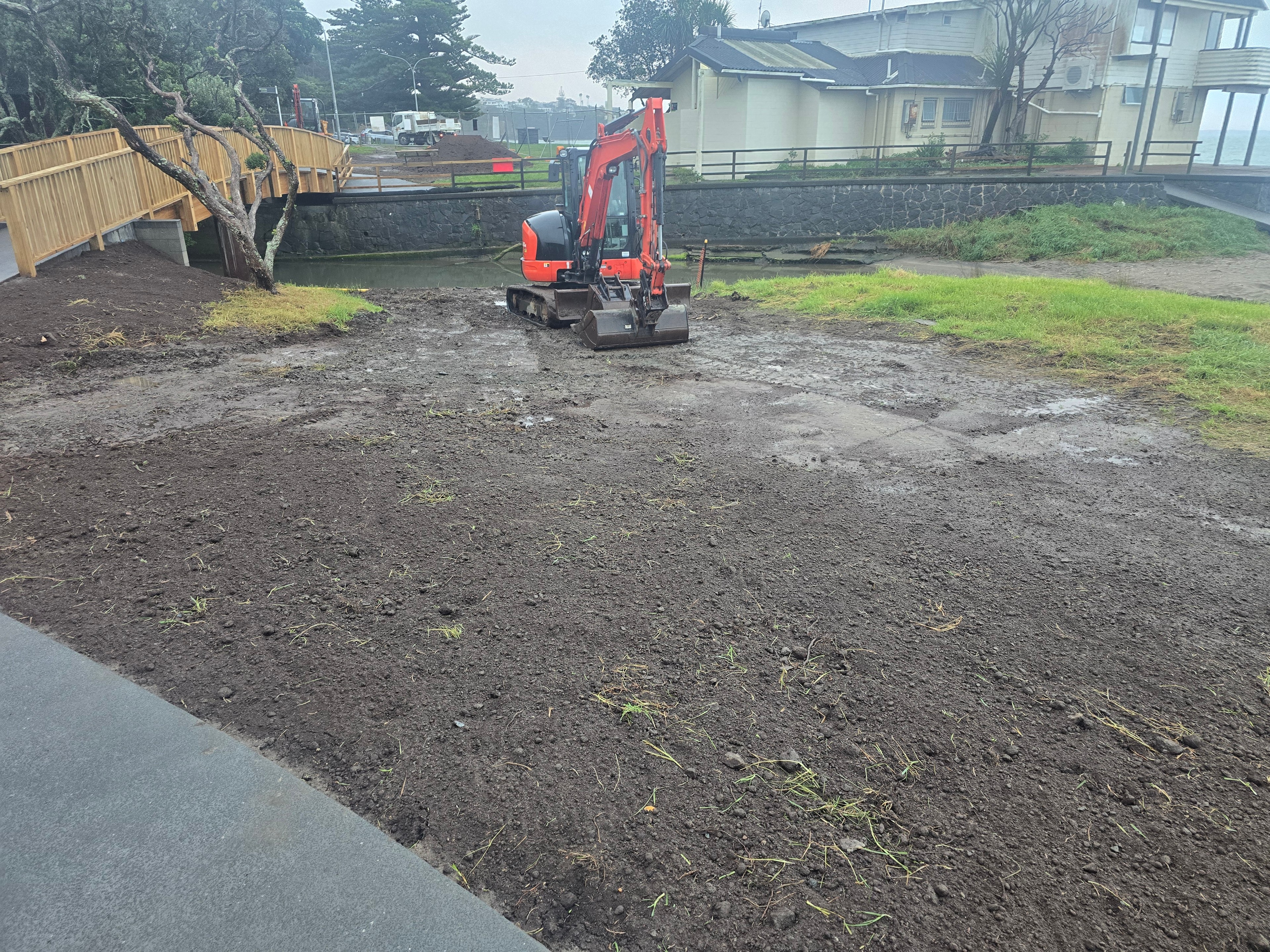Excavator standing on freshly spread topsoil beside a completed footbridge and stone wall