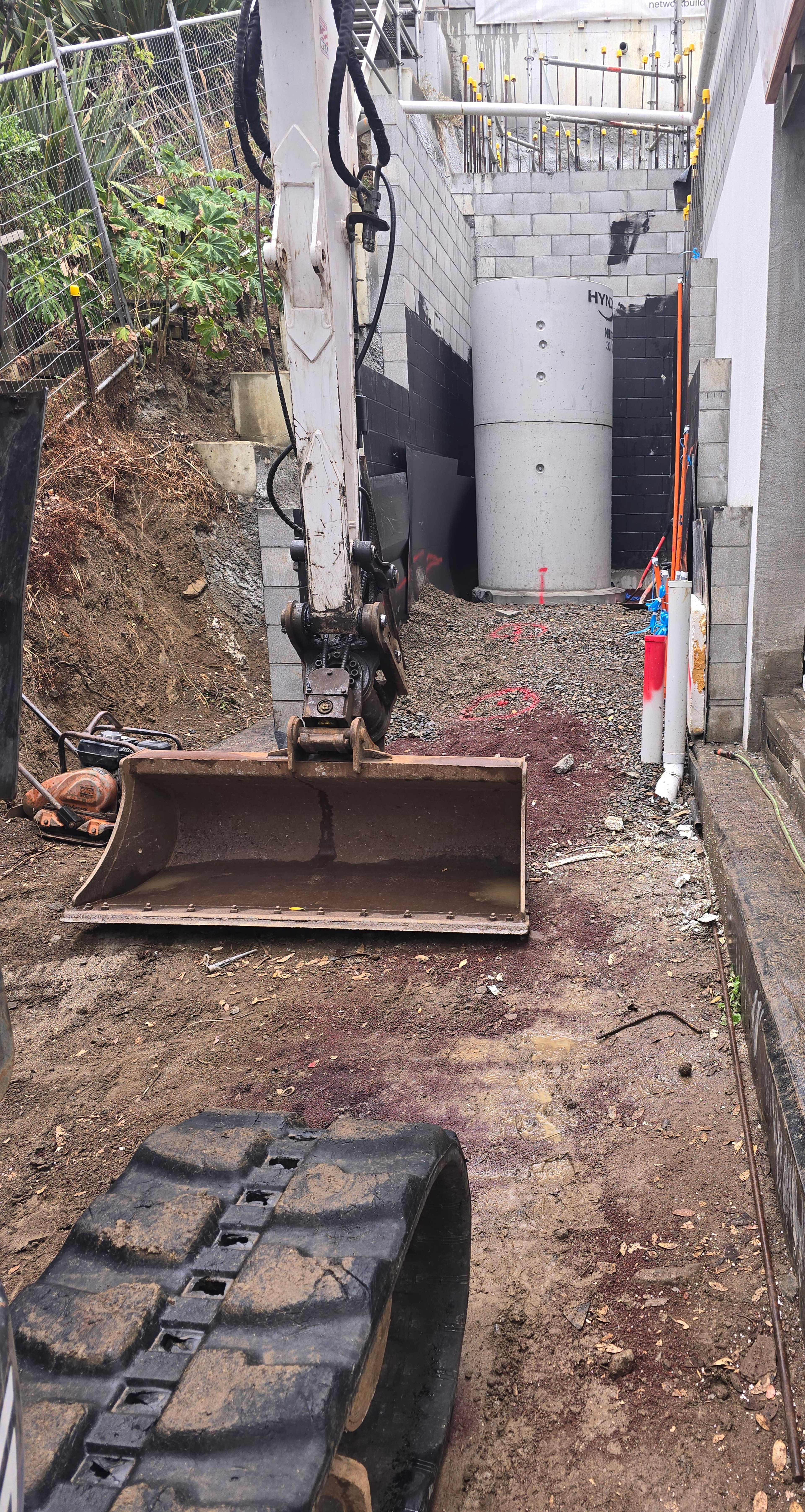 Excavator bucket at the base of a concrete manhole riser set between blockwork walls