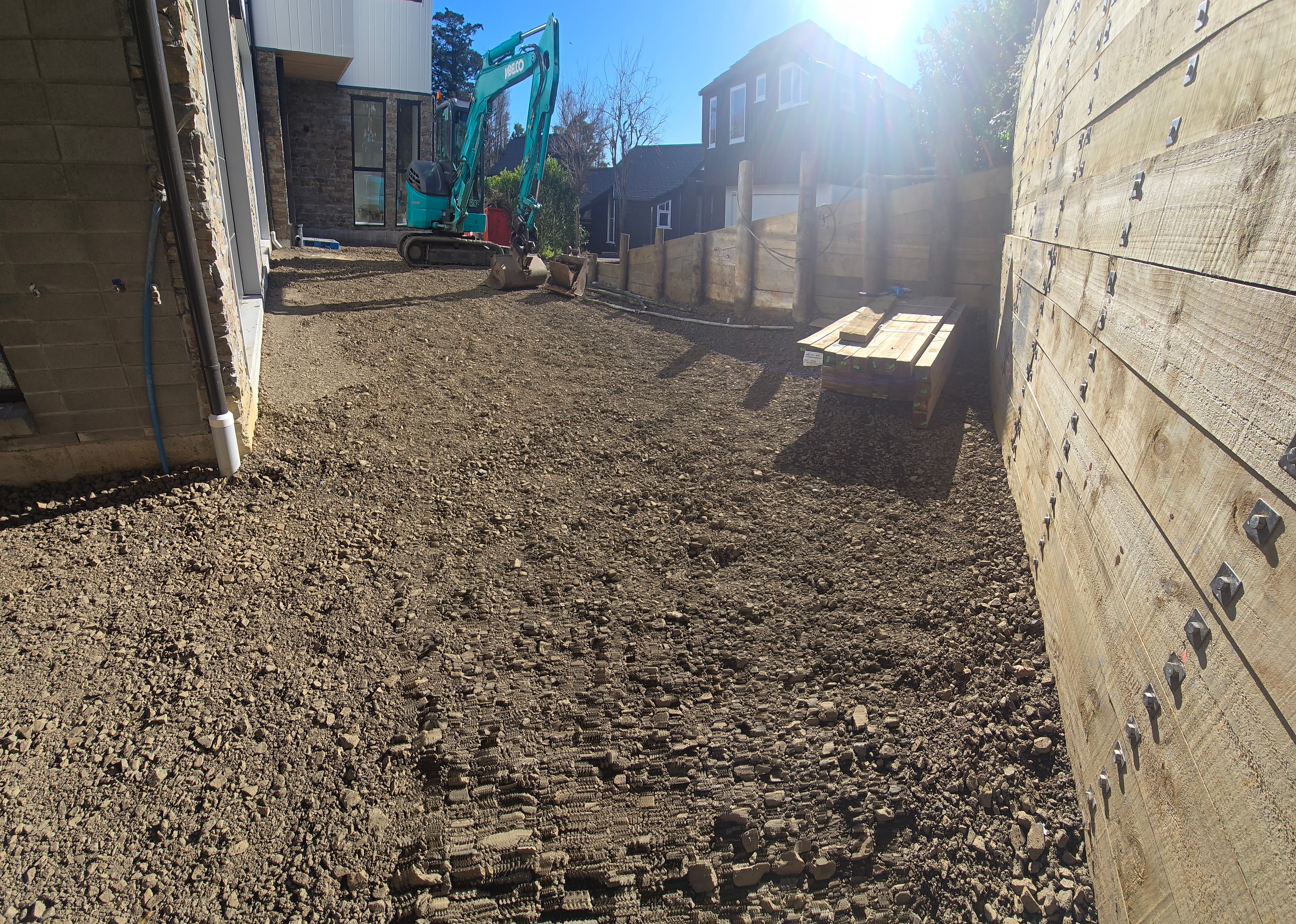 Timber retaining wall with steel plate fixings, backfilled with compacted basecourse, excavator alongside