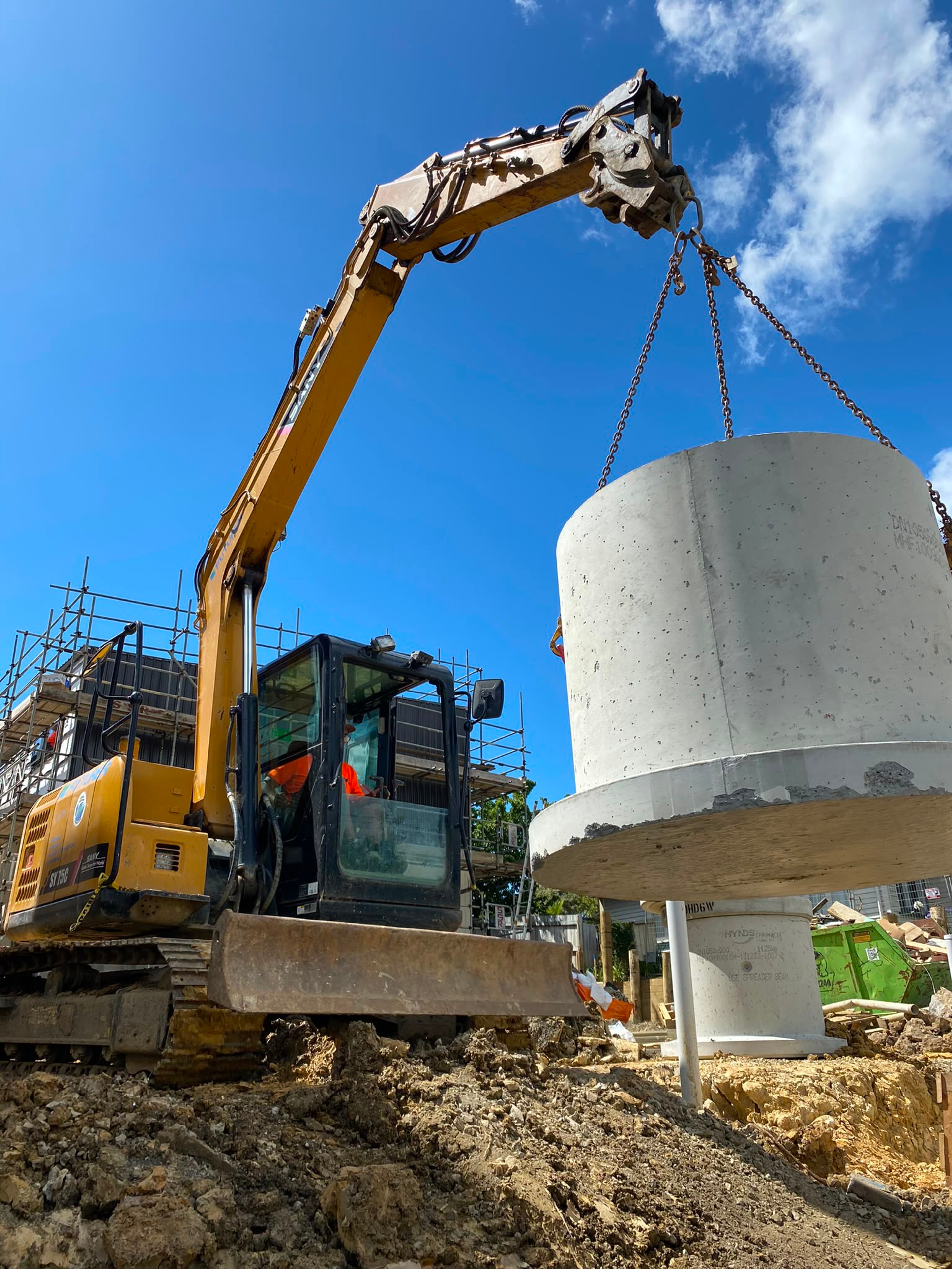 Excavator craning a concrete manhole chamber into position