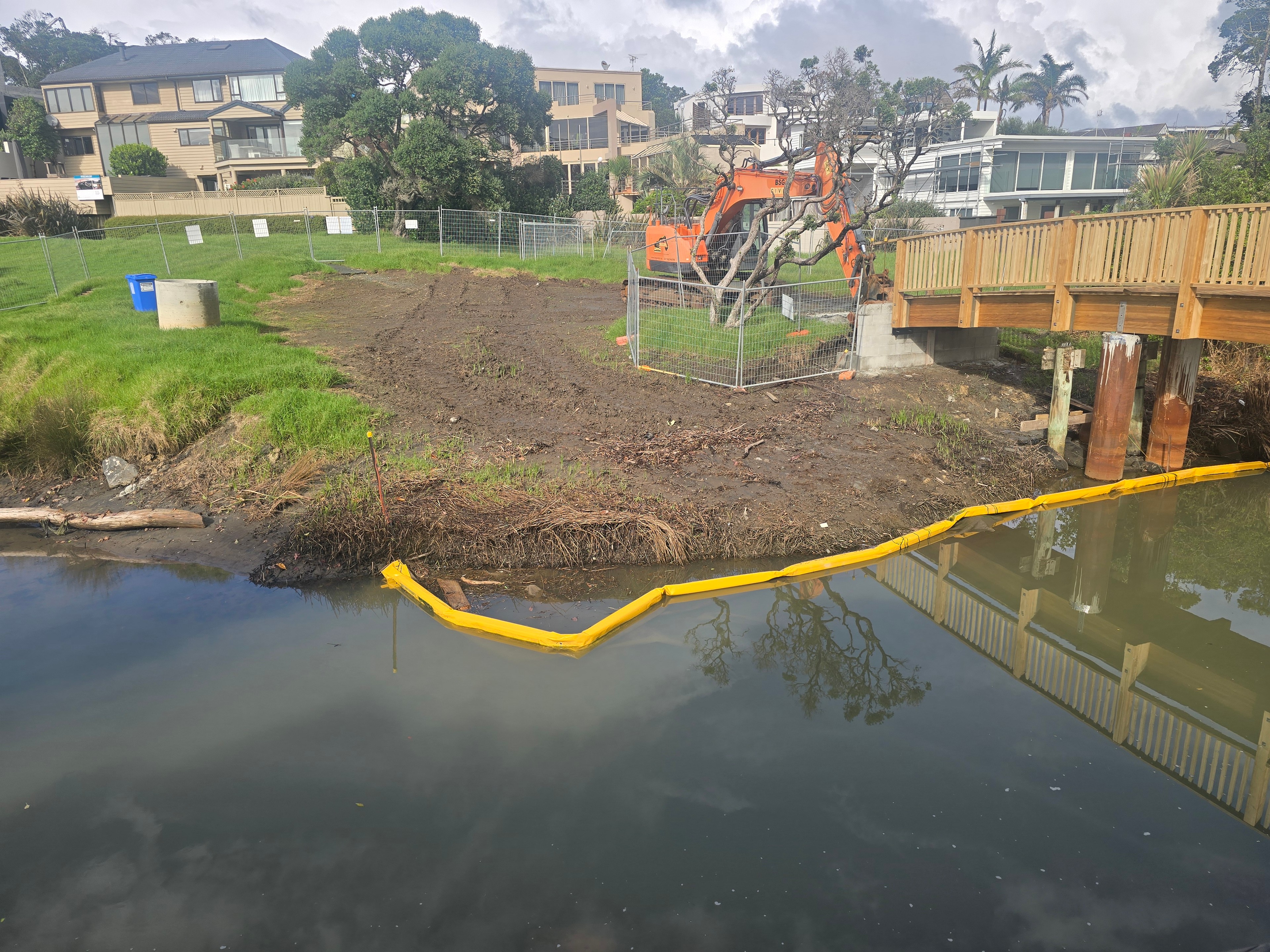 Excavator working behind temporary fencing beside a new timber footbridge, with a silt boom laid across the estuary