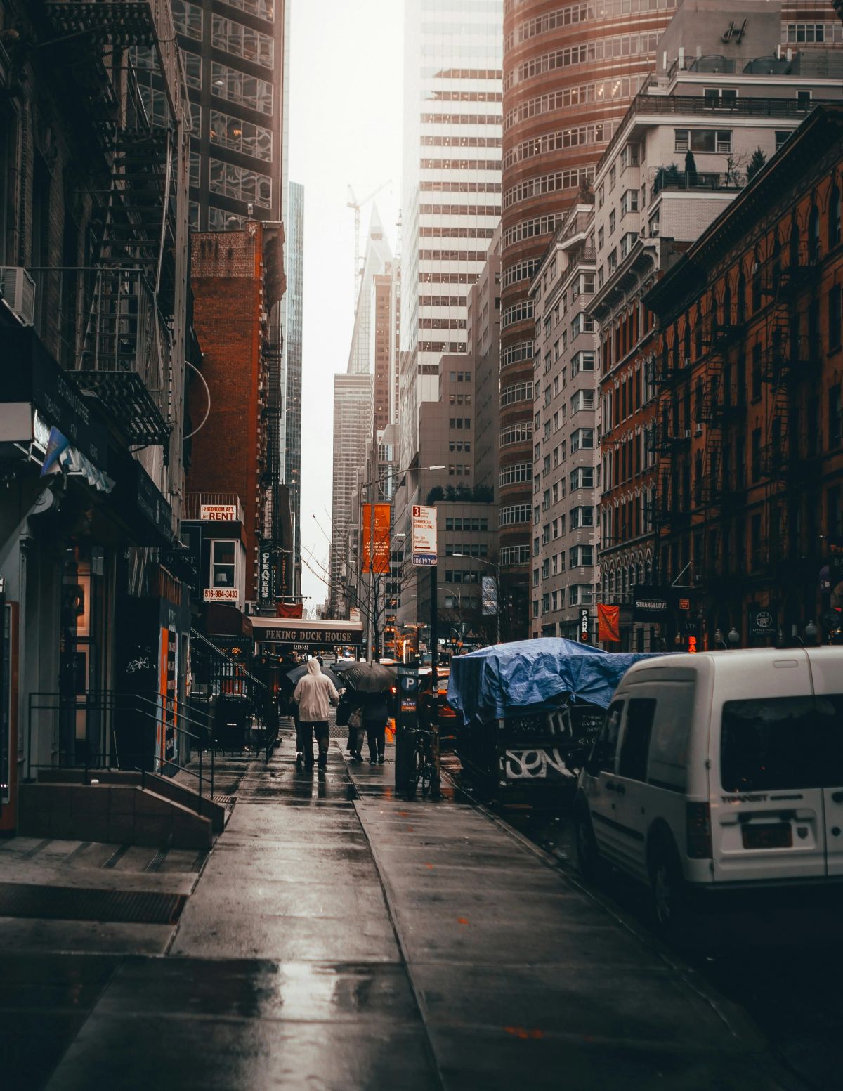 A wet Midtown sidewalk between brick walk-ups and fire escapes, a white van parked at the curb