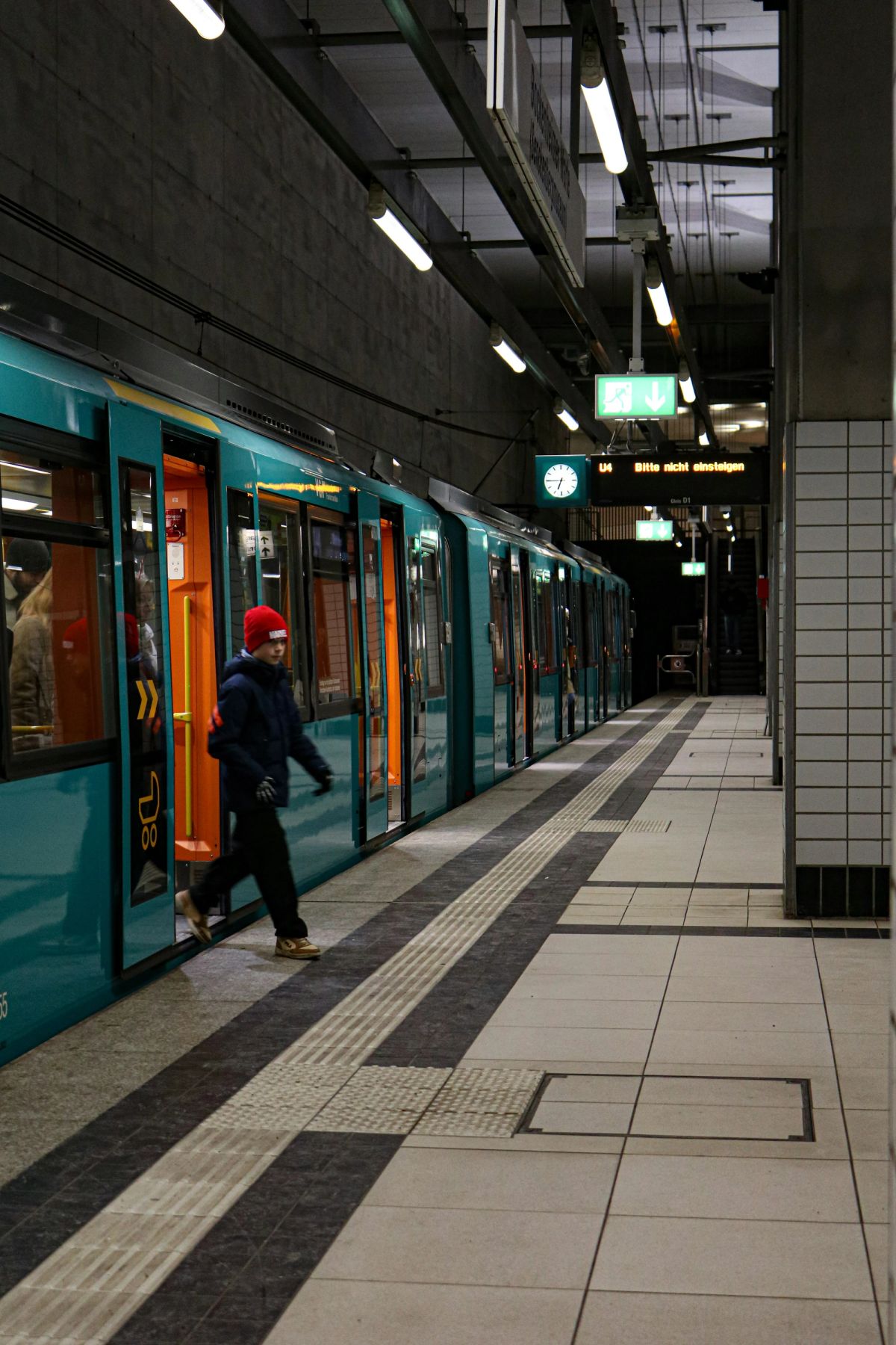 A man in a red beanie steps off a teal train onto a tiled platform