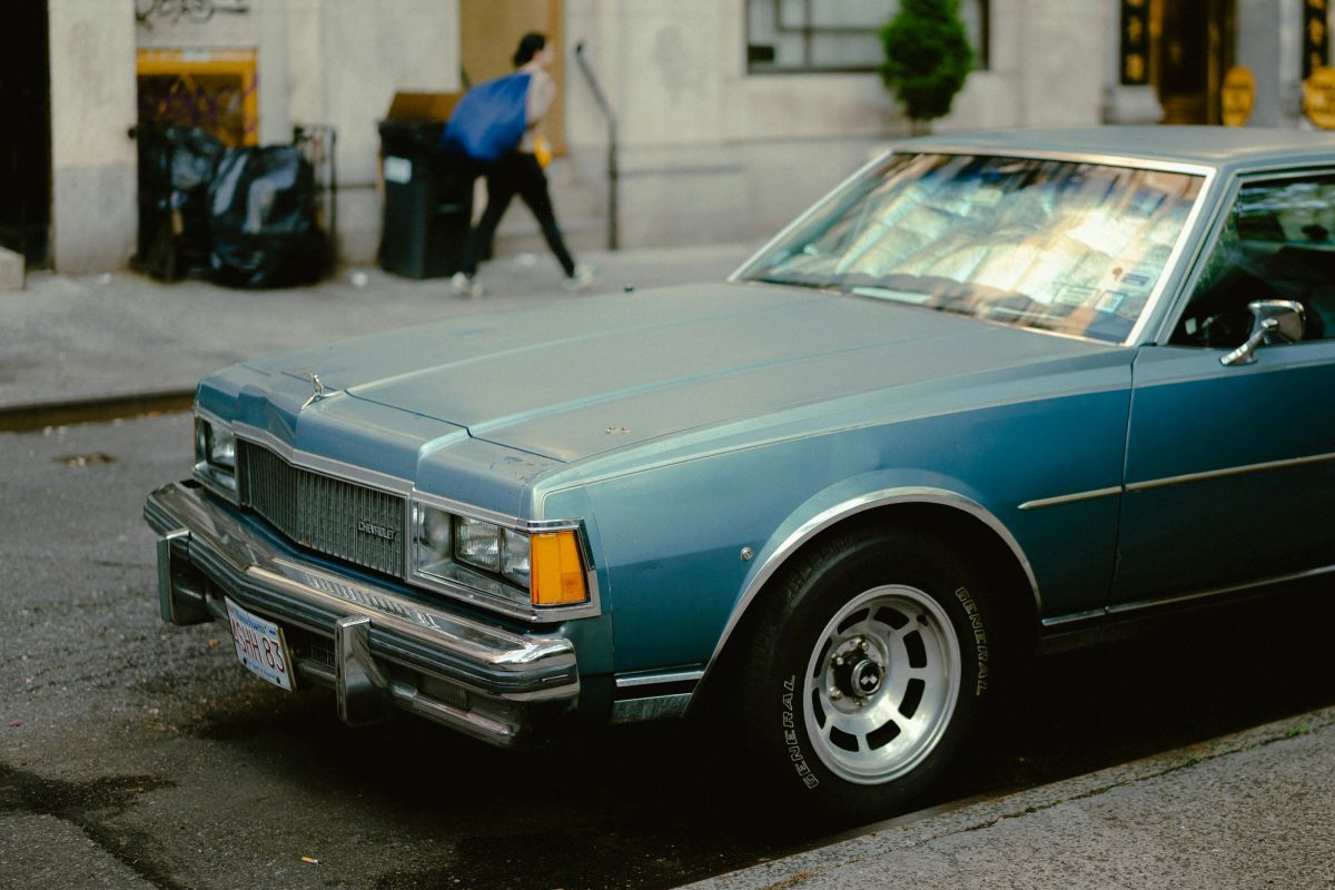 A pale blue vintage sedan parked at the curb as a woman walks past with a blue bag