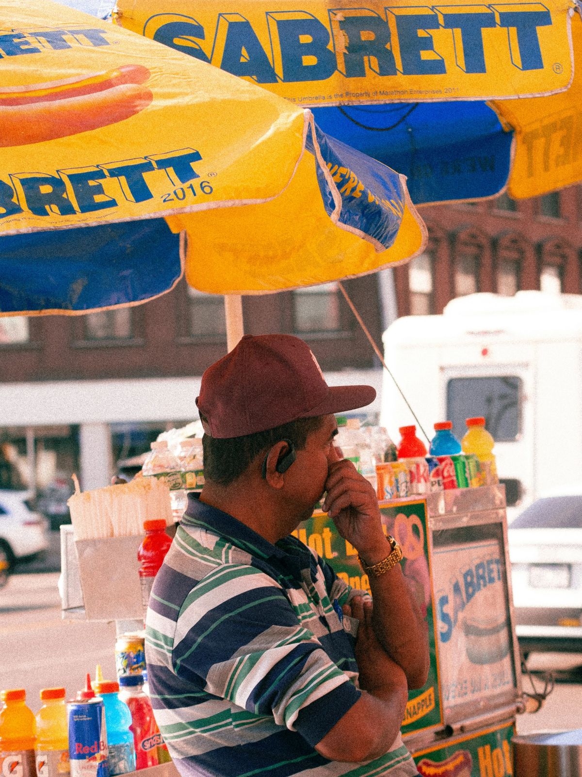 A hot dog vendor in a maroon cap under a yellow and blue Sabrett umbrella