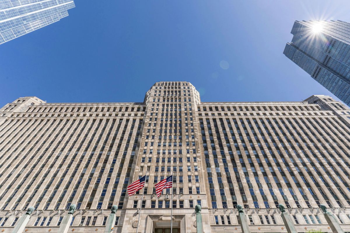 Looking up at a pale stone Art Deco tower with American flags over the entrance