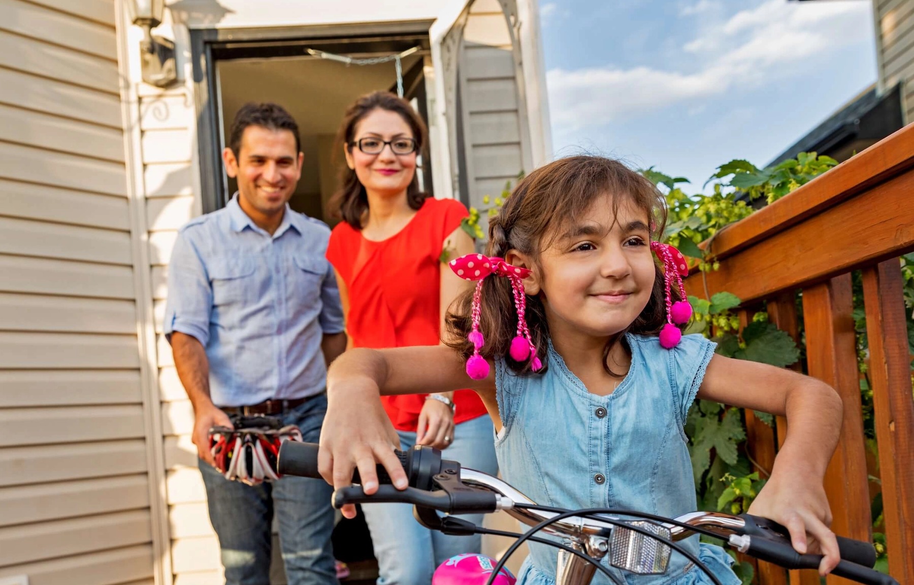 Two parents follow behind a young girl with pink decorations in her hair as she pushes a white bike along their home's deck.