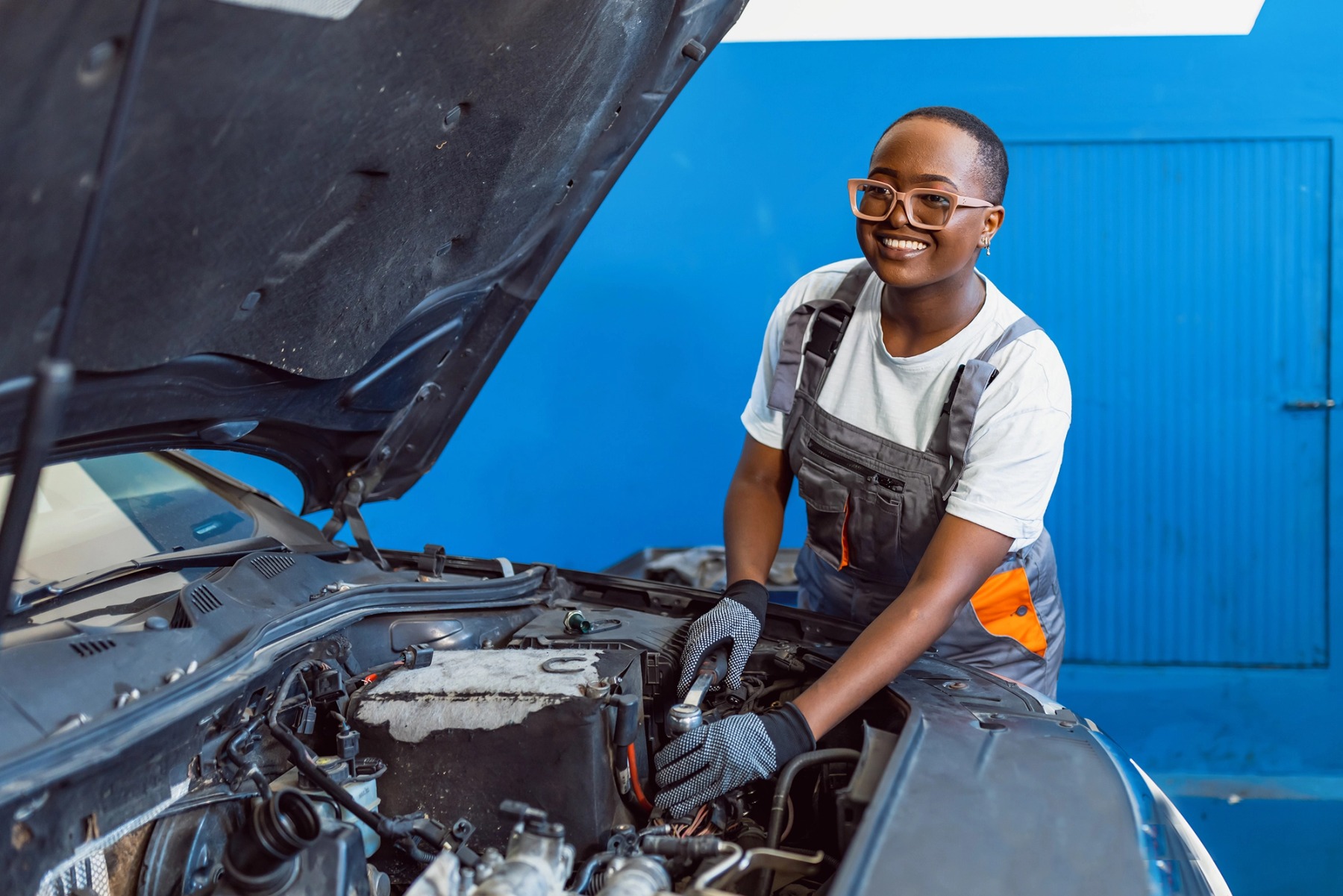 A person wearing glasses and overalls, with shaved head and earrings, works on a vehicle engine.