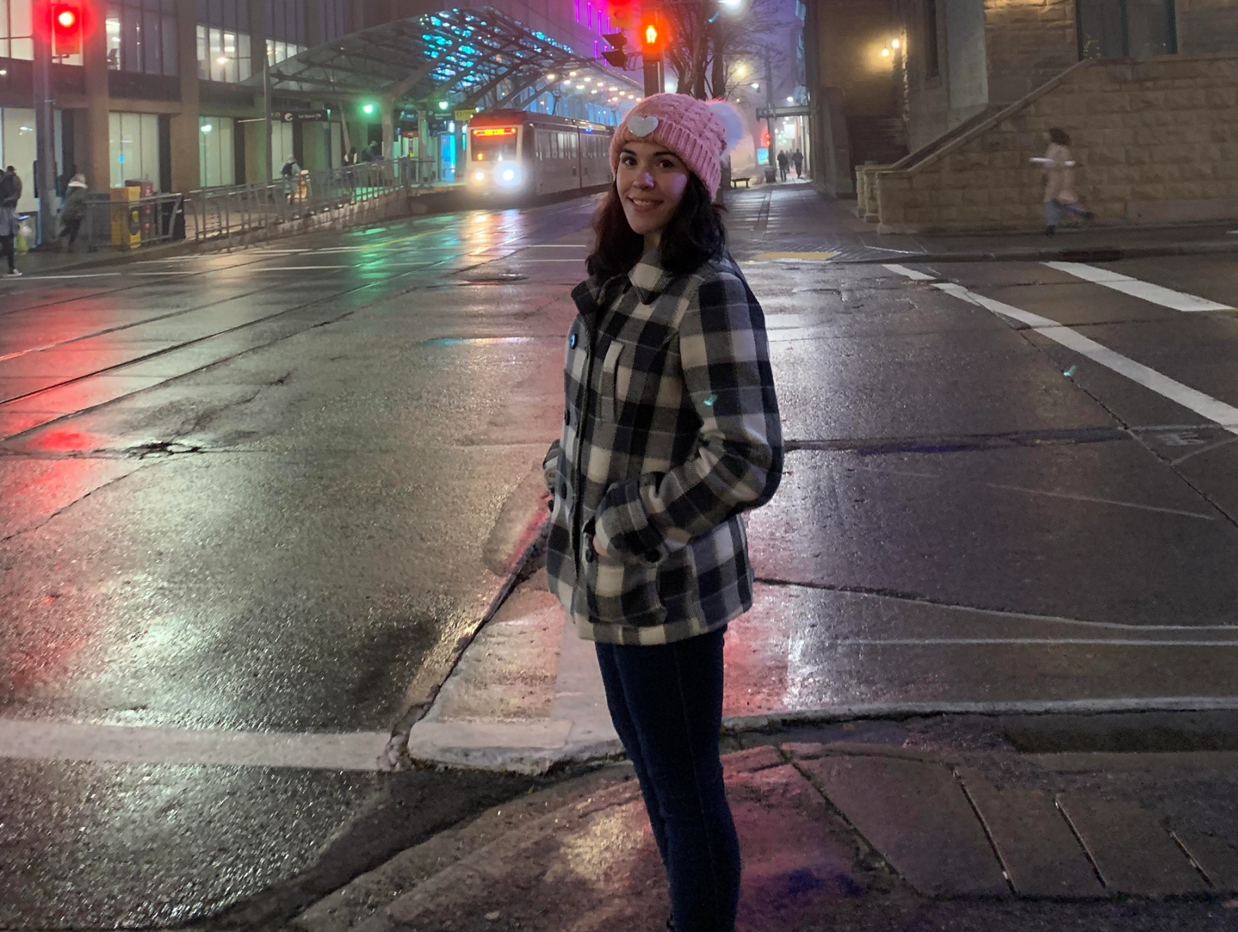 A young woman wearing a plaid coat and pink toque stands on a corner in downtown Calgary, smiling at the camera.