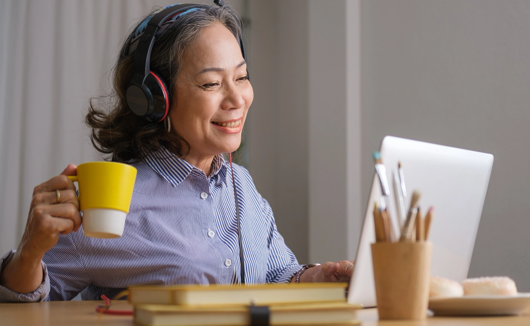 An older woman sitting at a desk holding a mug and looking at her laptop.