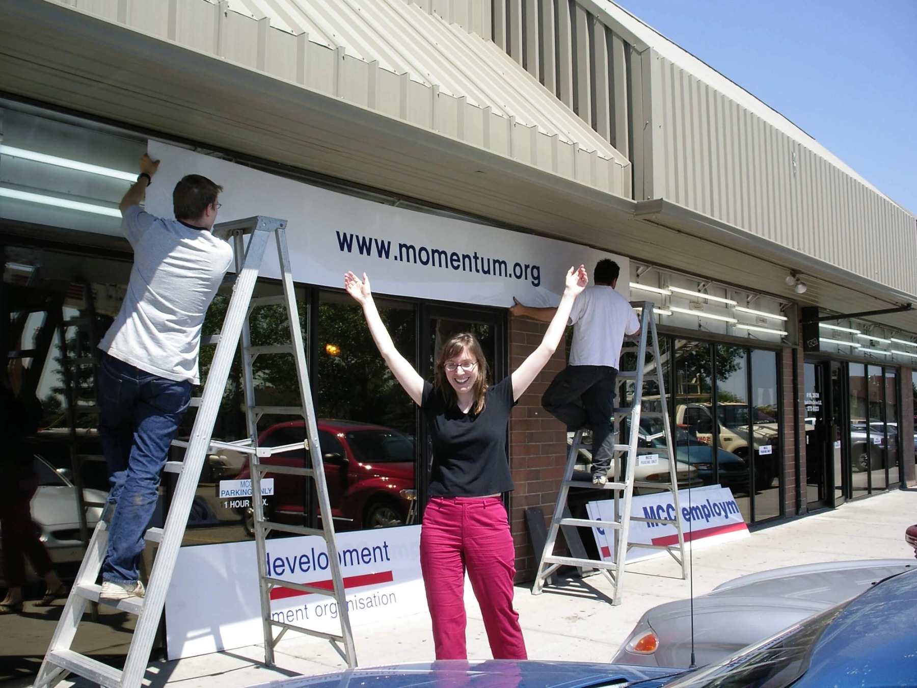 A woman in a black shirt and pink pants smiles and waves as two signage workers hang a sign reading "www.momentum.org" on the building behind her.