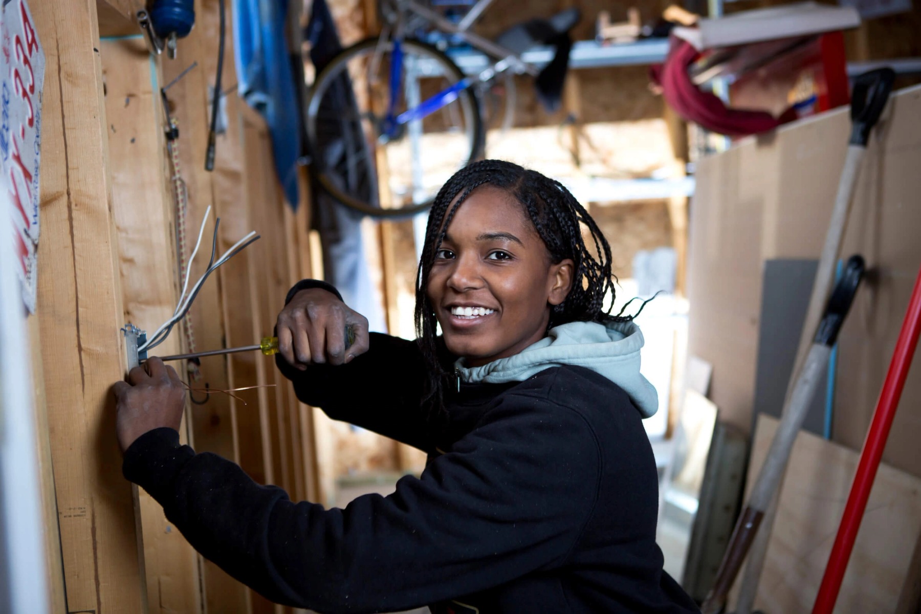 A woman in a black sweater handles electrical wiring along an unfinished wall.