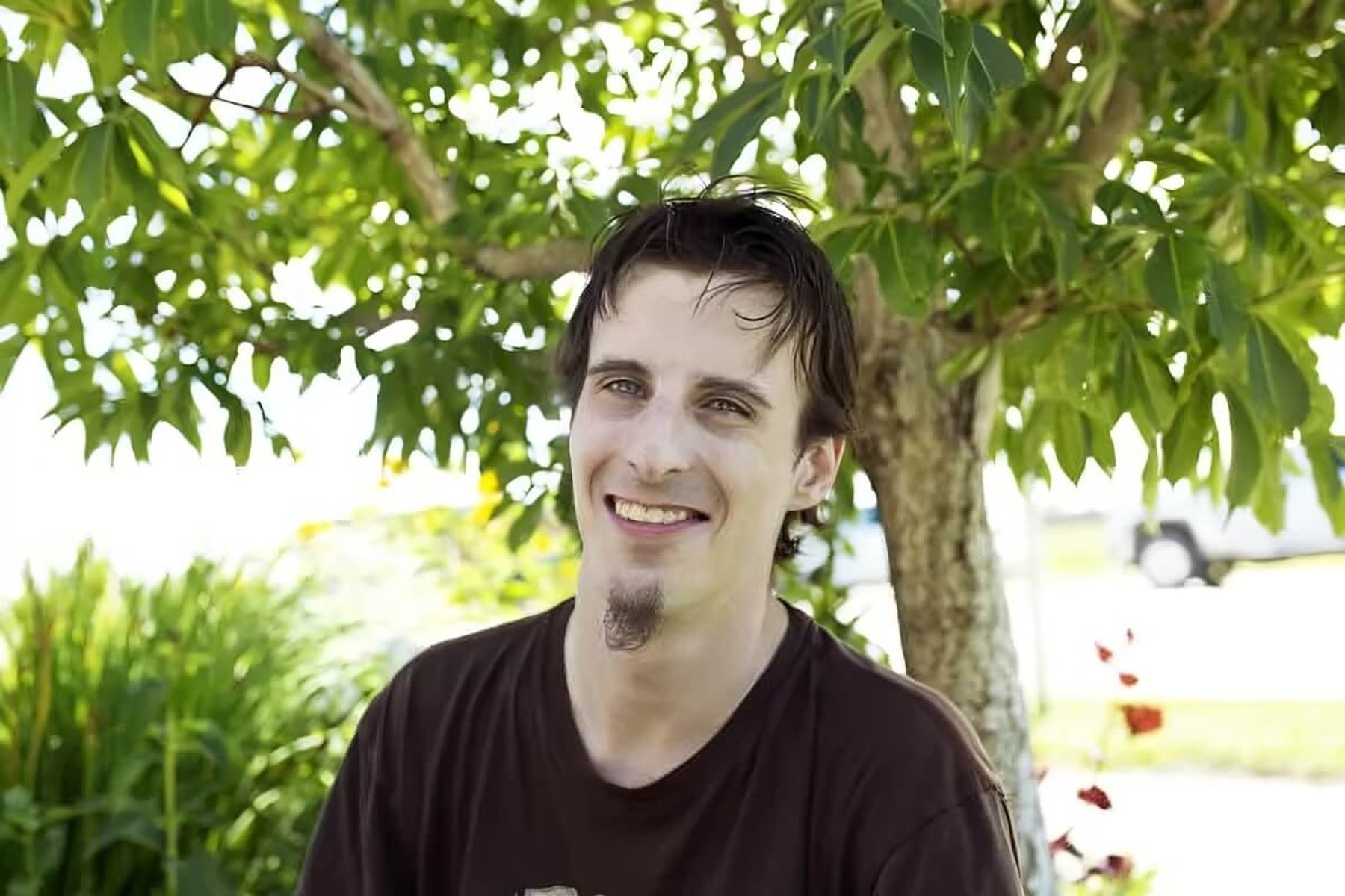 A man with a goatee smiles, standing in front of a green tree.