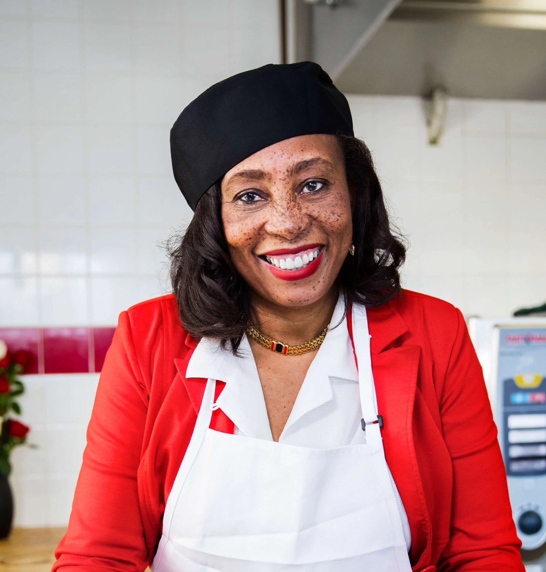 Head and shoulders portrait of a black woman with brown hair, wearing a black baker's hat and white apron, smiling and looking at the camera.