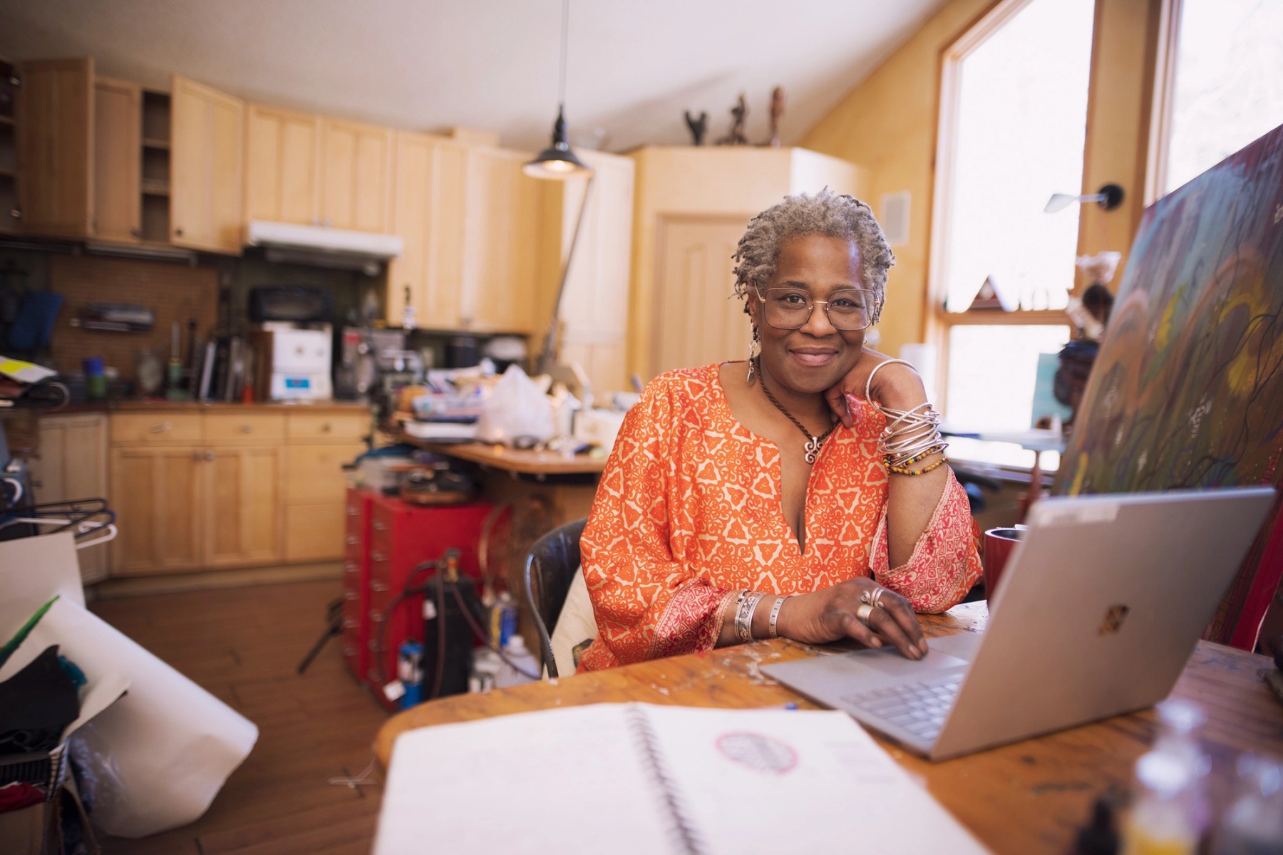 A black woman with grey hair and glasses sits at a table in her art studio, smiling at the camera.