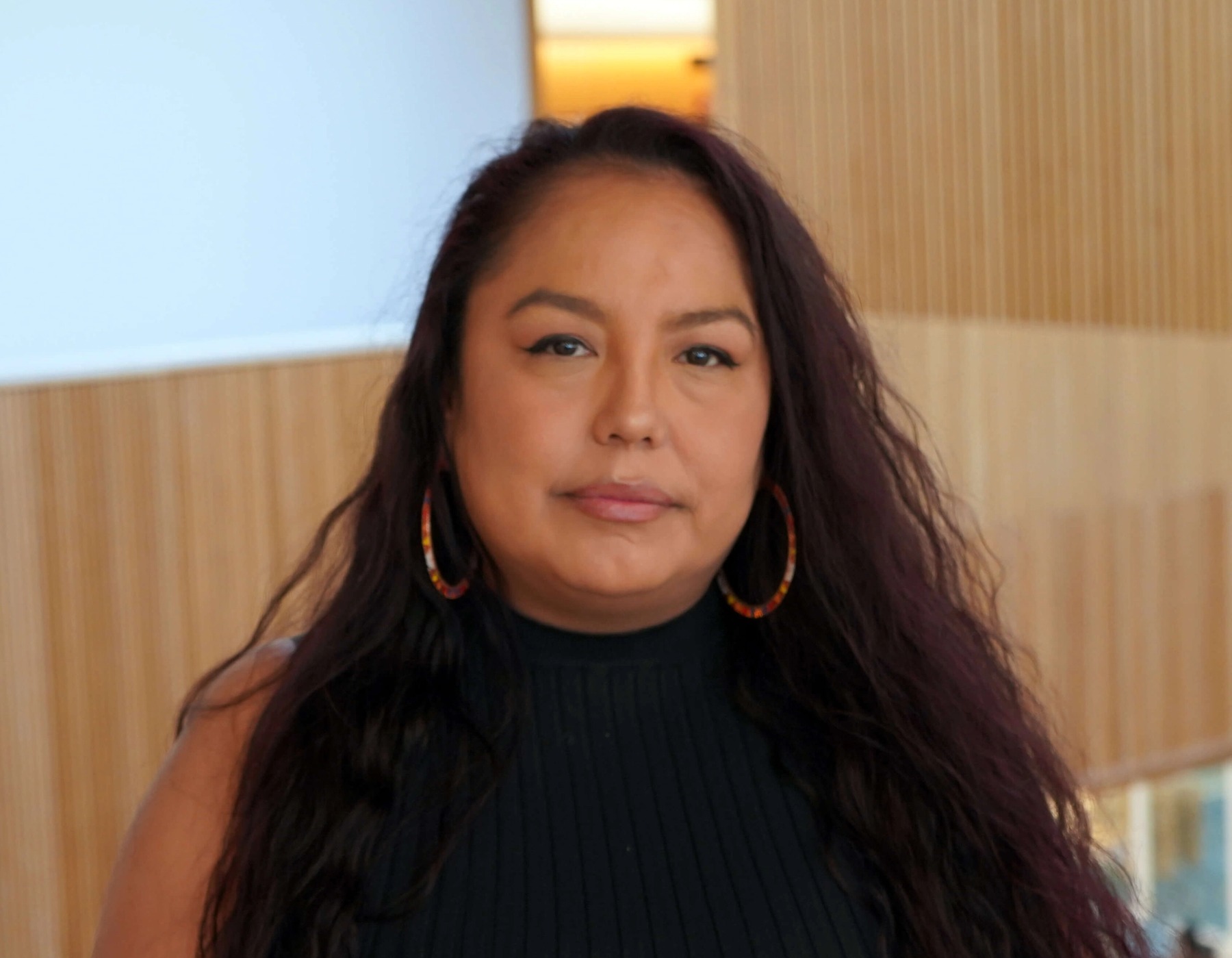 Head and shoulders portrait of an Indigenous woman with long dark hair, a black top and hoop earrings.