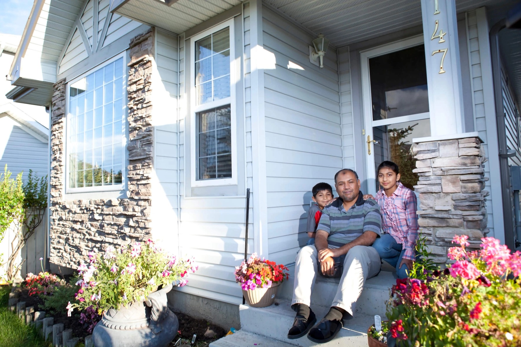A father and two children sit in front of a suburban home, surrounded by thriving flowers.