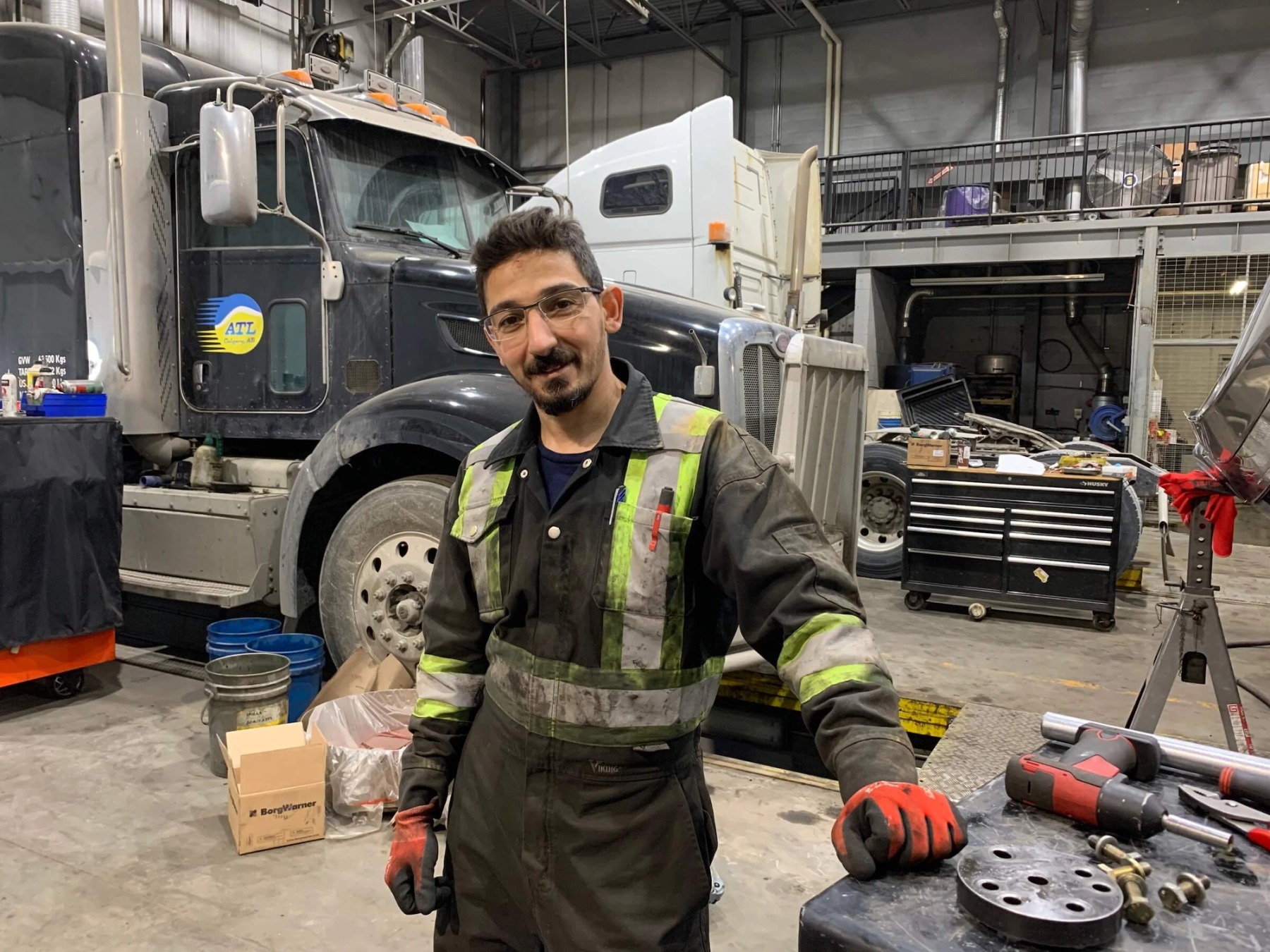 A young person in black coveralls and safety gear poses for a photo in an auto shop, in front of a large black semi truck.