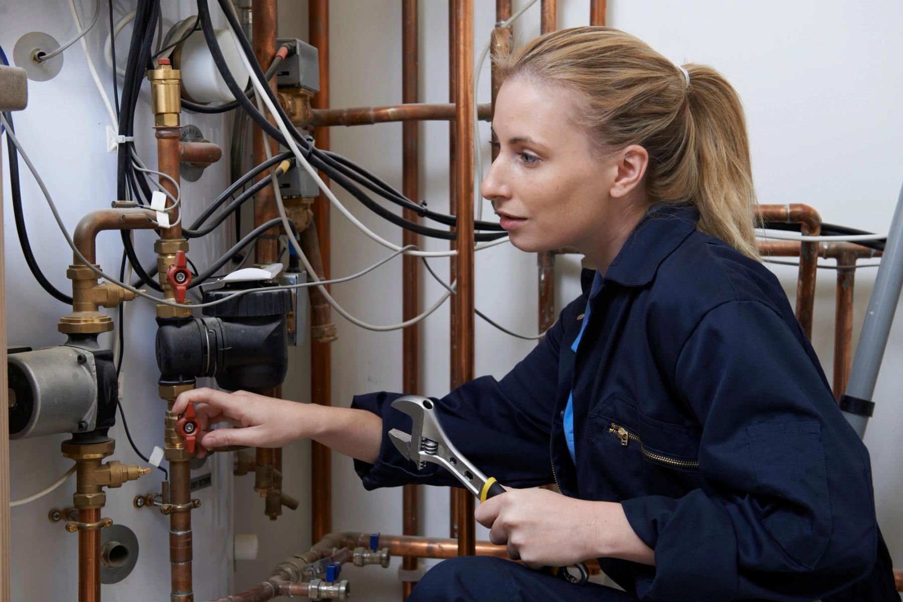 A woman with blonde hair is holding a wrench and working on plumbing pipes.