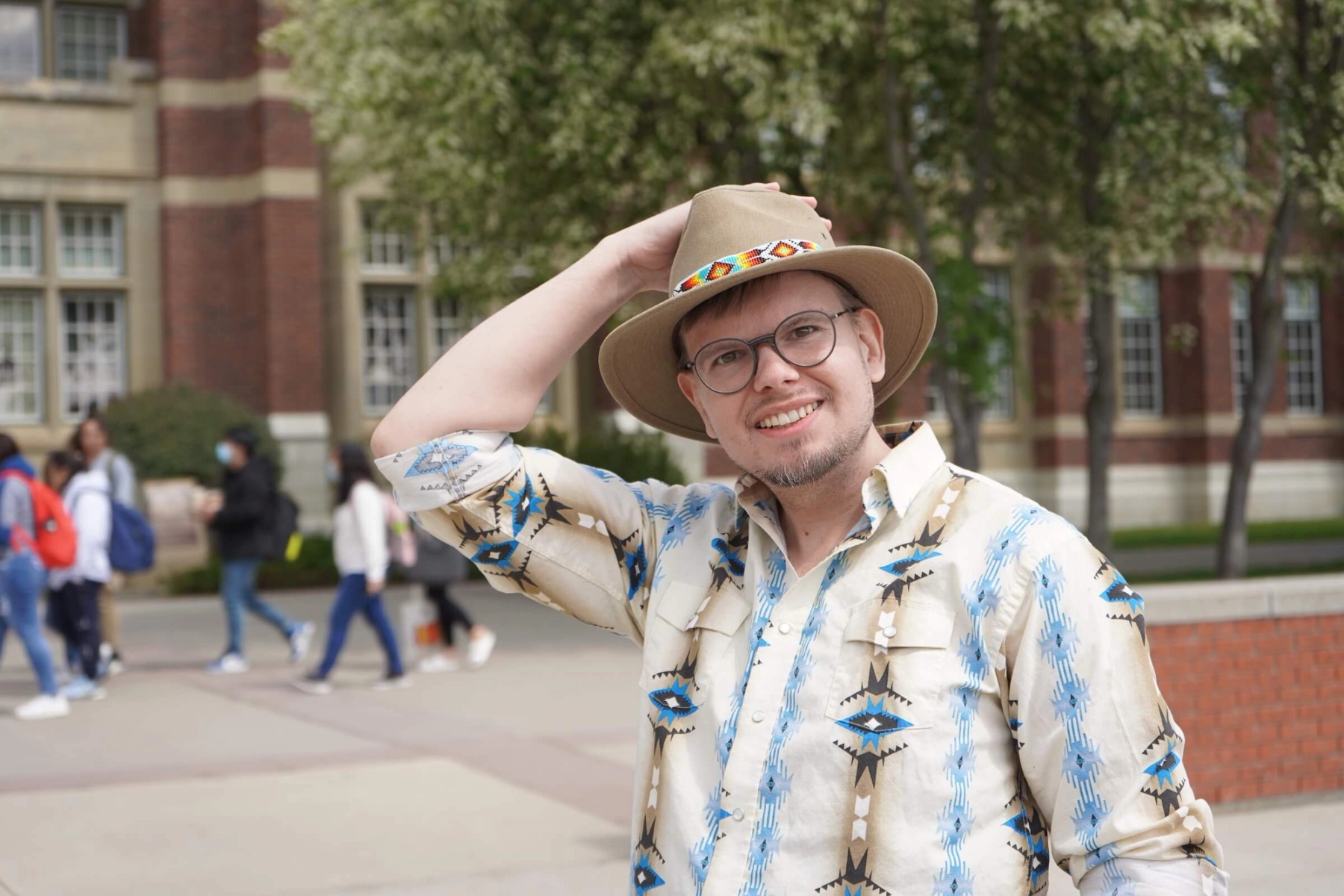 A young man in rounded glasses and a patterned shirt and hat poses outside a school.