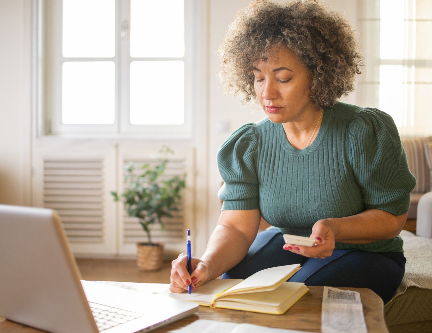 A woman sits in her living room with a laptop open, she is writing in a notebook.