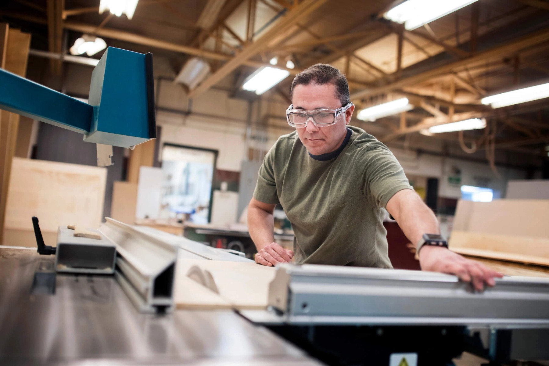 A man in a green shirt and safety goggles uses a large saw in a carpentry workshop.