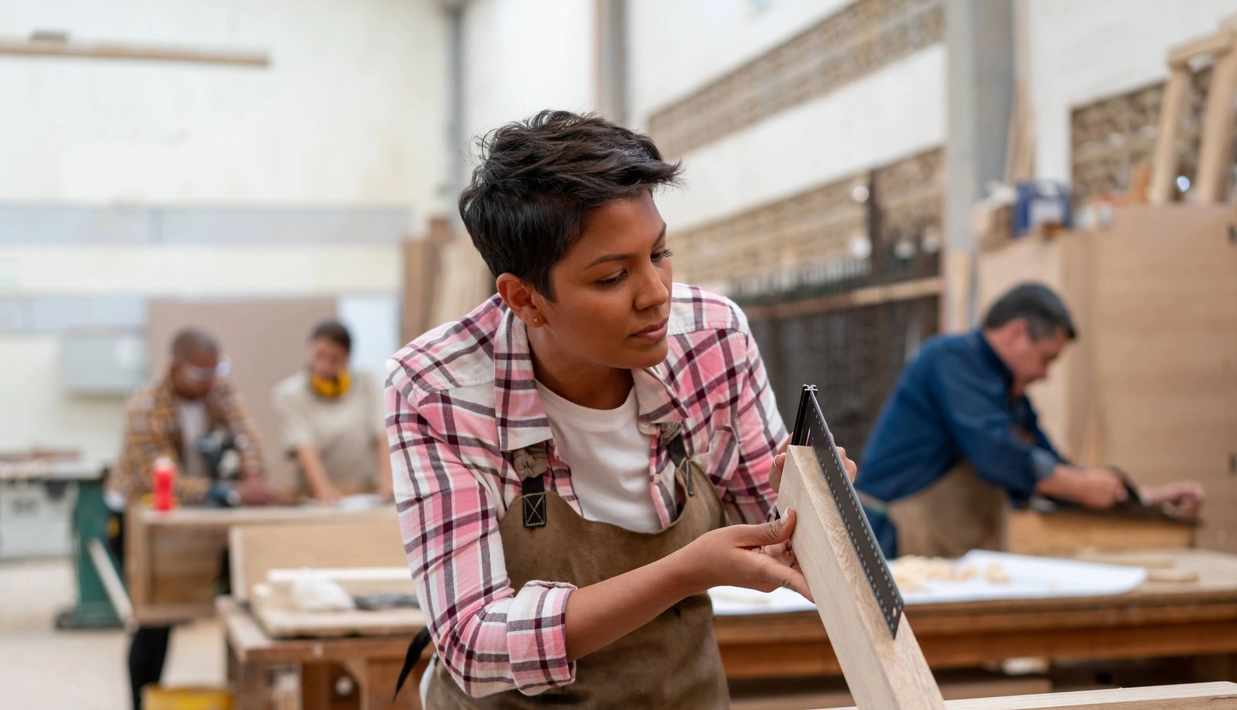 A person with short hair and a pink plaid shirt measures a piece of wood in a carpentry workshop.