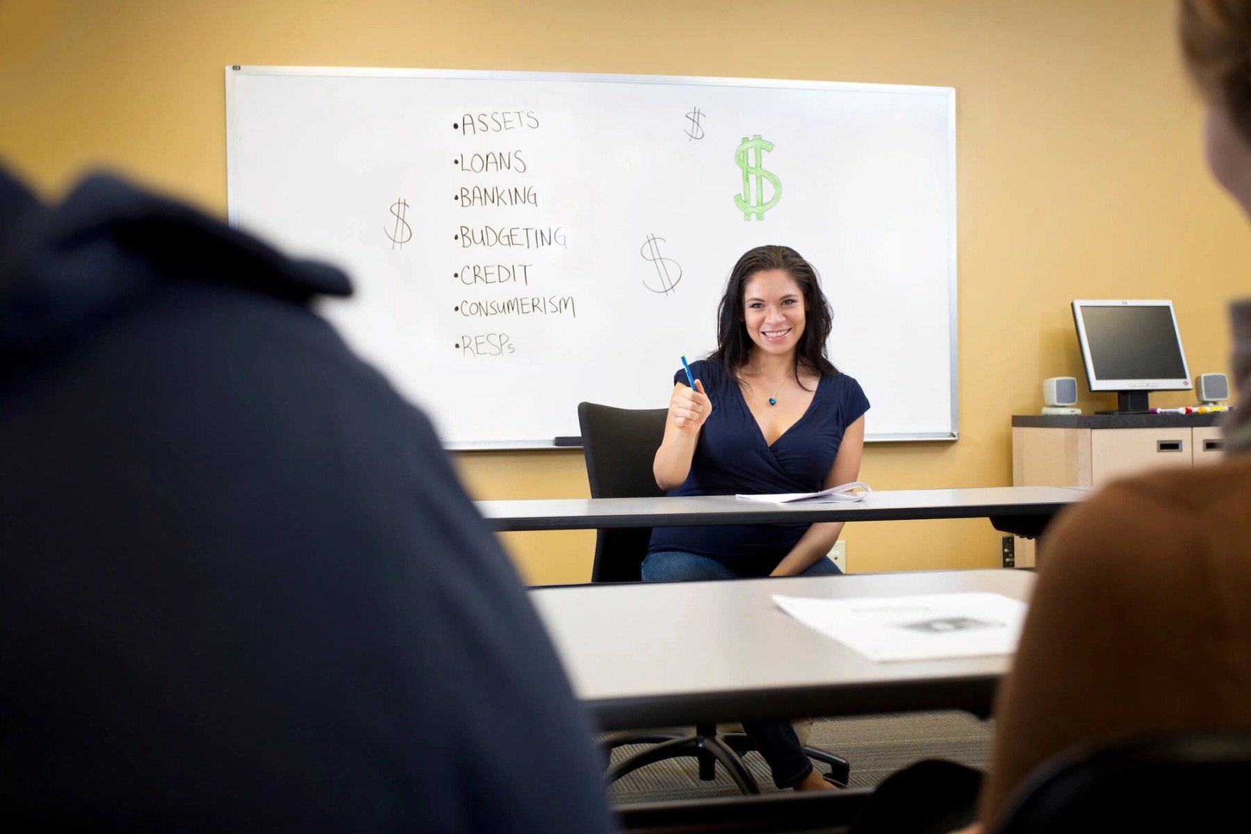 A woman addresses questions in front of a whiteboard reading 'Assets, Loans, Banking, Budgeting, Credit, Consumerism, RESPs'.