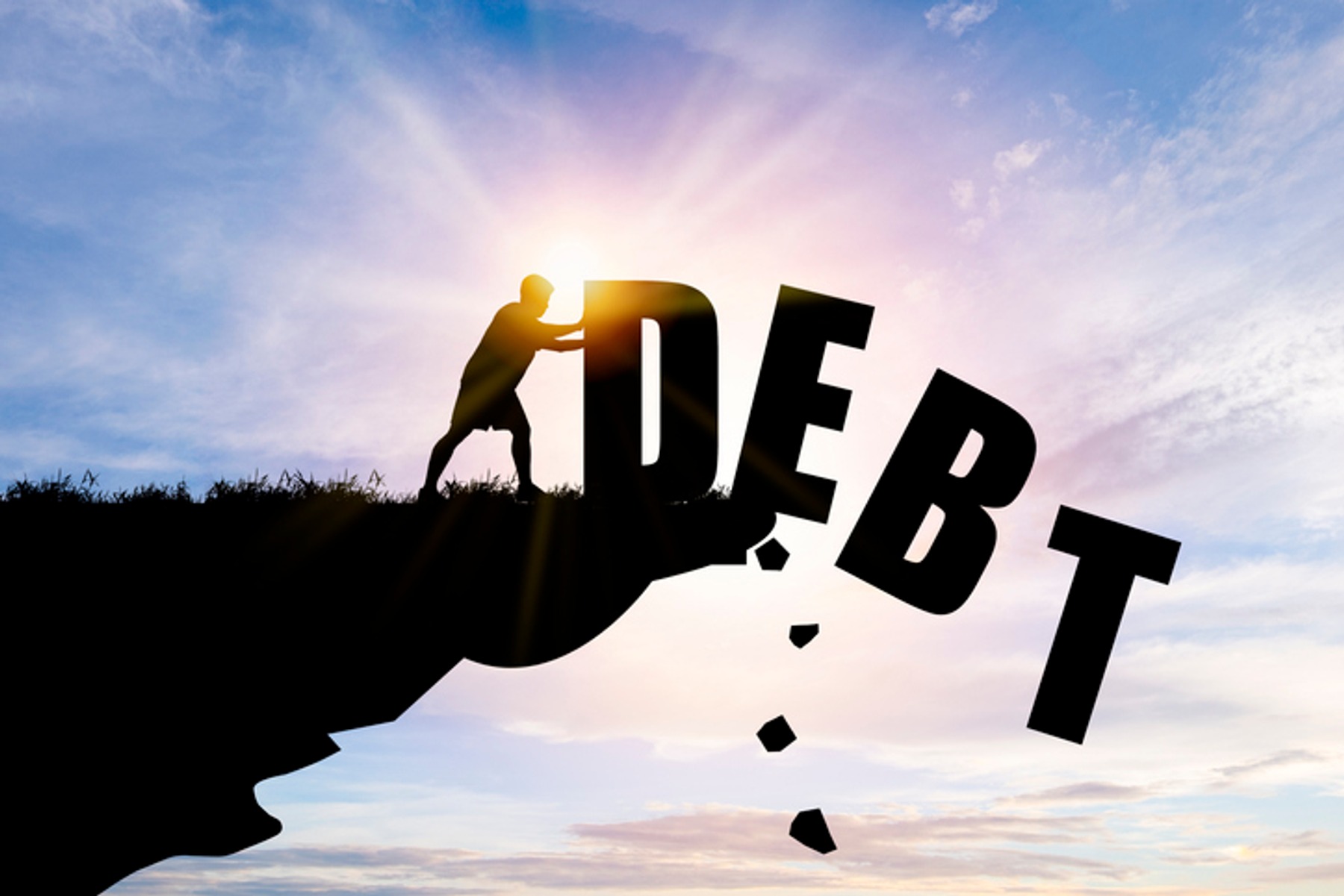 Silhouette view of a person pushing giant letters that spell "debt" off a cliff.