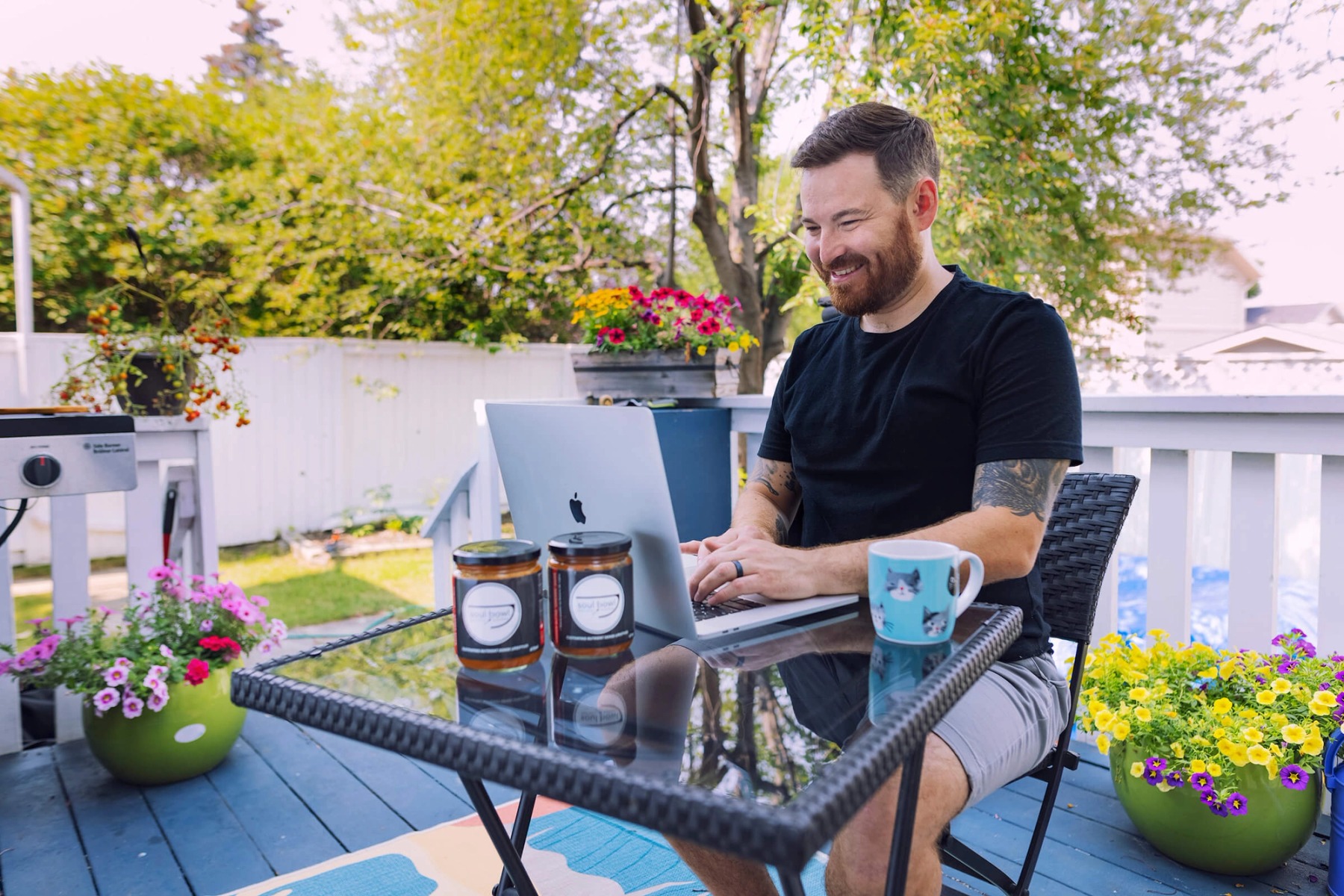 A man with a black shirt and red beard types on a laptop at a patio table in a backyard, surrounded by flower pots.