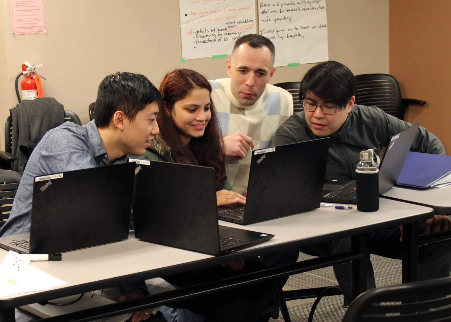 Four people in a classroom surround a black laptop for a group project.