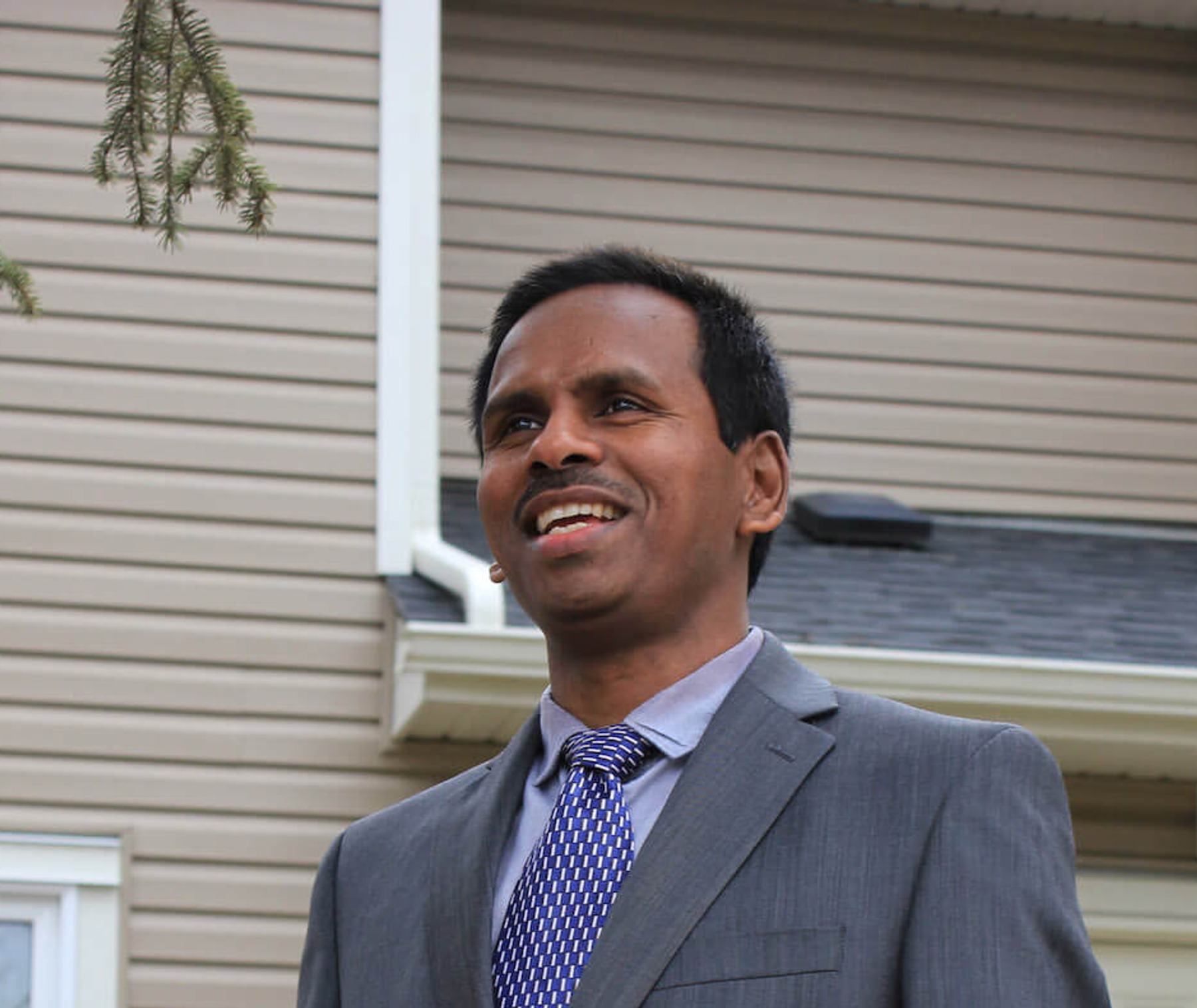 A man in a grey suit and blue tie poses for a photo in front of a brown building.