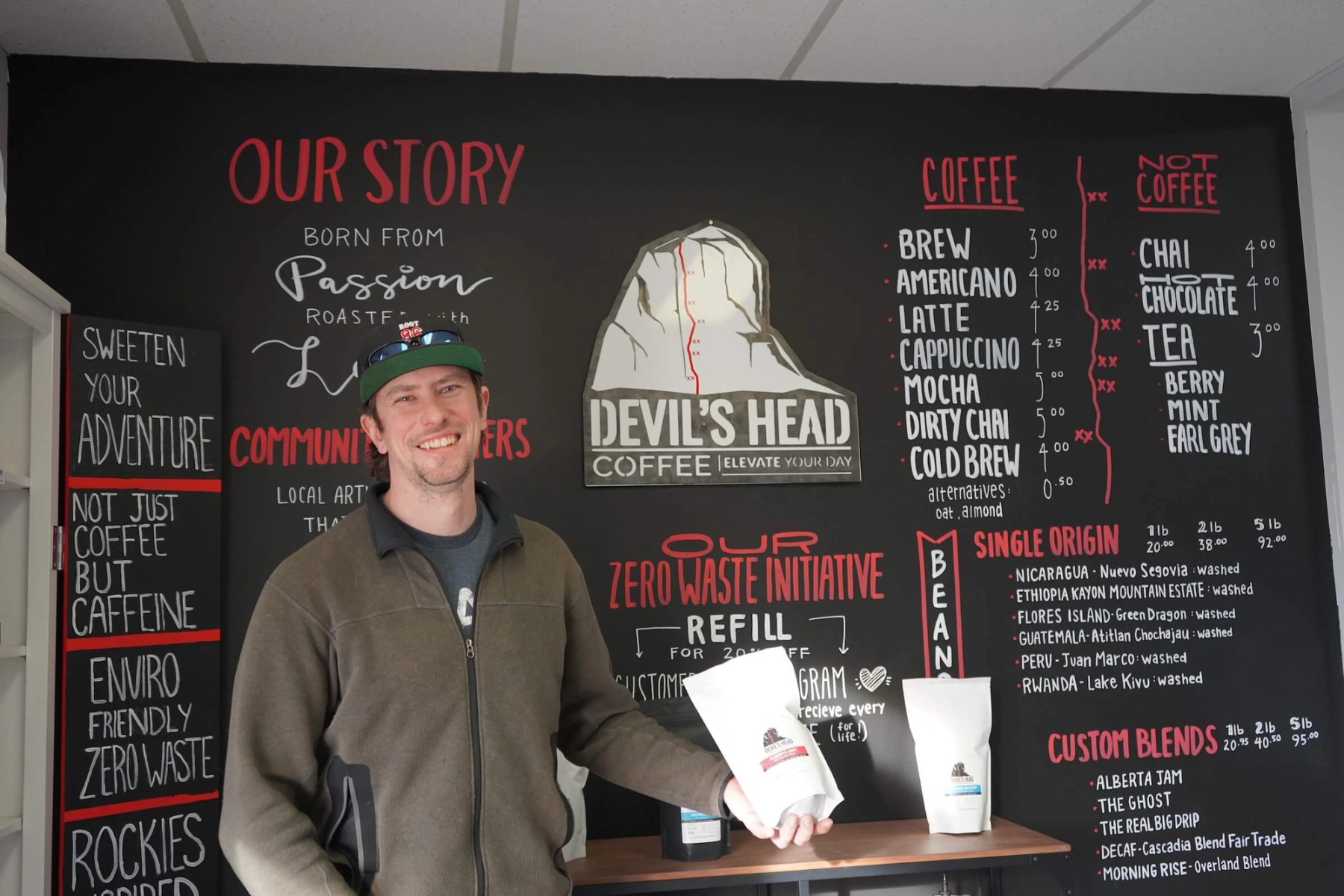 A man in a baseball hat and brown sweater poses in front of a menu board reading 'Devil's Head Coffee' with an assortment of menu items and their respective prices.