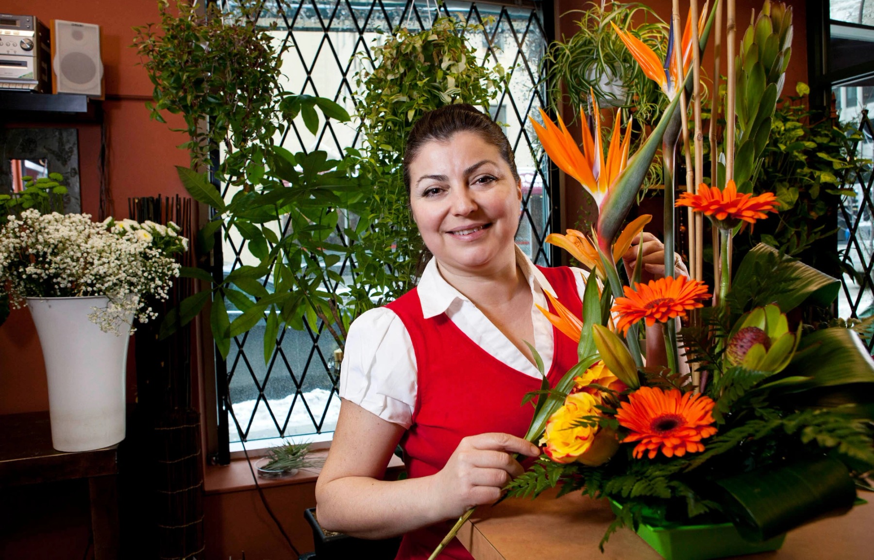 A woman wearing a white shirt and red vest poses behind an arrangement of orange and yellow flowers in a warmly-lit store.