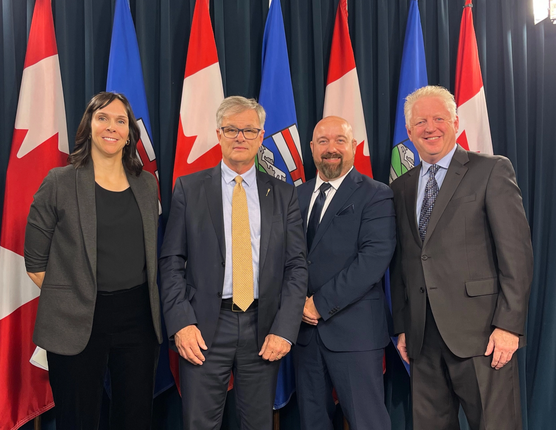Four people standing side by side in front of legislative flags