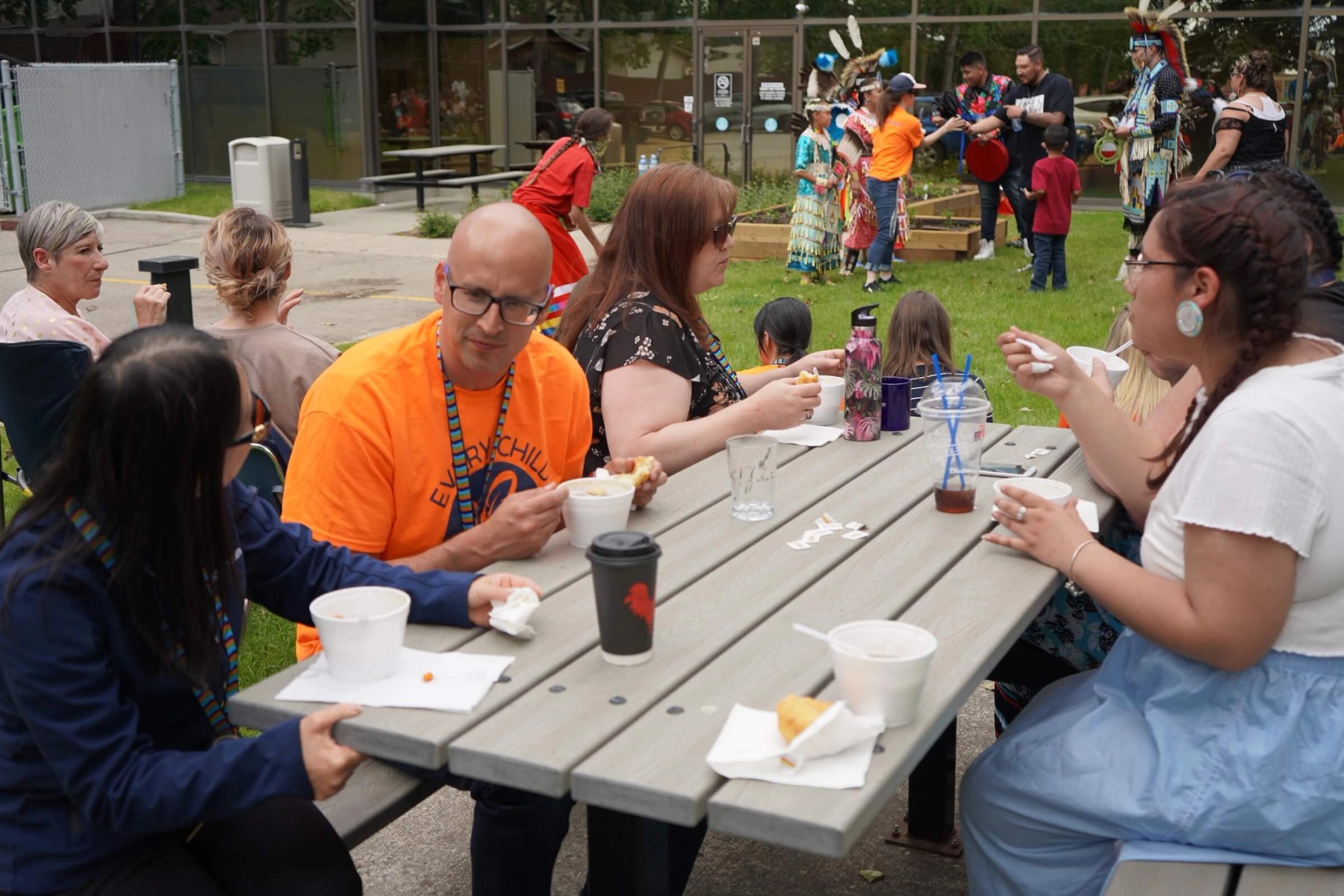 A group of people chat at a grey picnic table while eating food and drinking coffee.