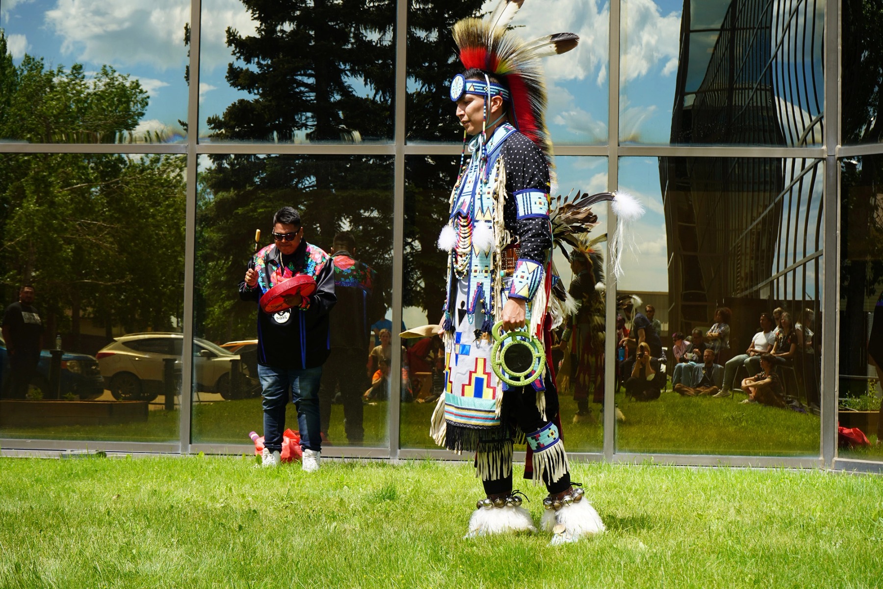 An Indigenous dancer stands outdoors on green grass, while a singer drums and sings. A reflective office building wall is in the background showing an audience.