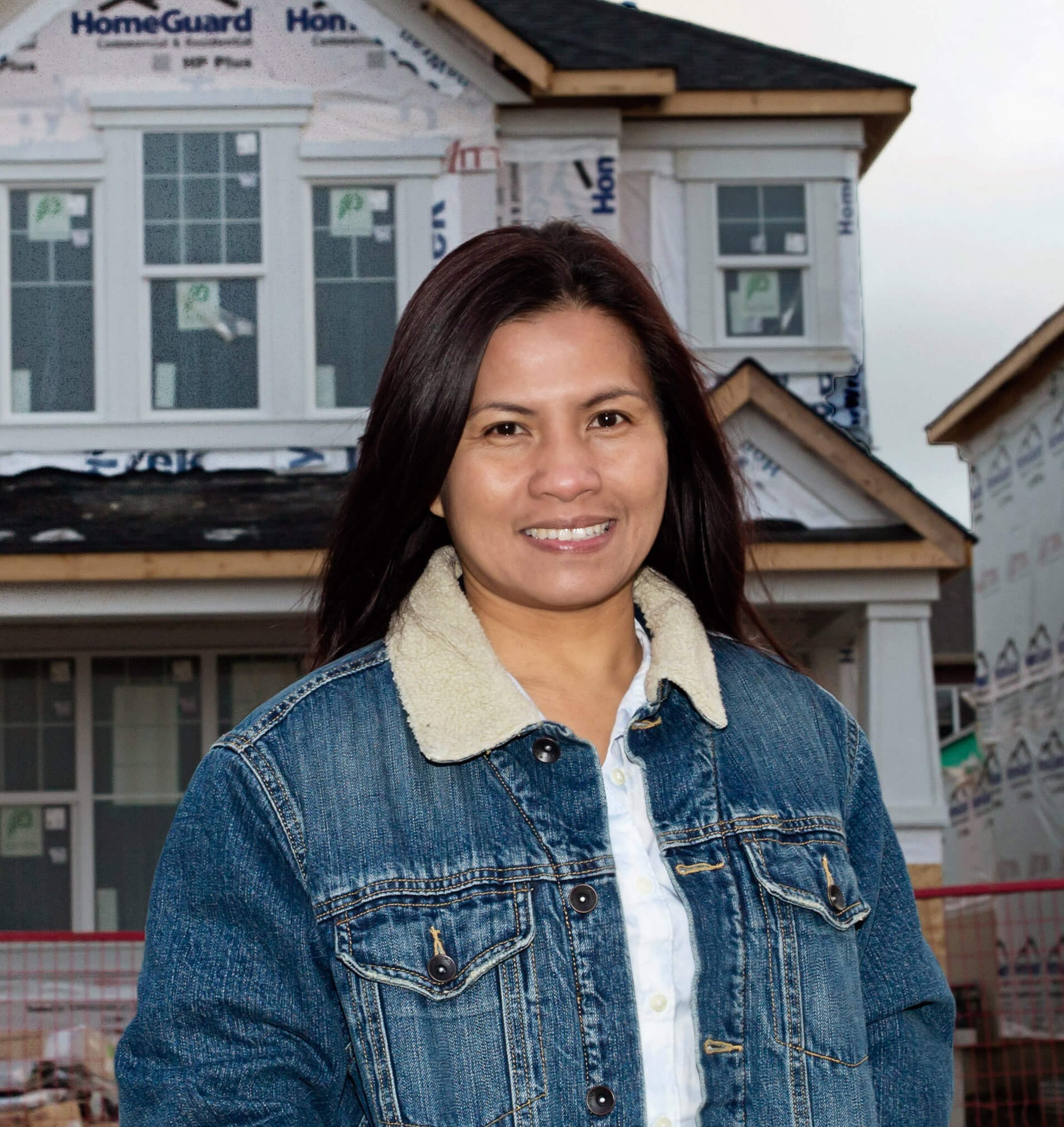 Head and shoulders portrait of a woman of Asian descent, standing in front of a row of homes being built, smiling at the camera.