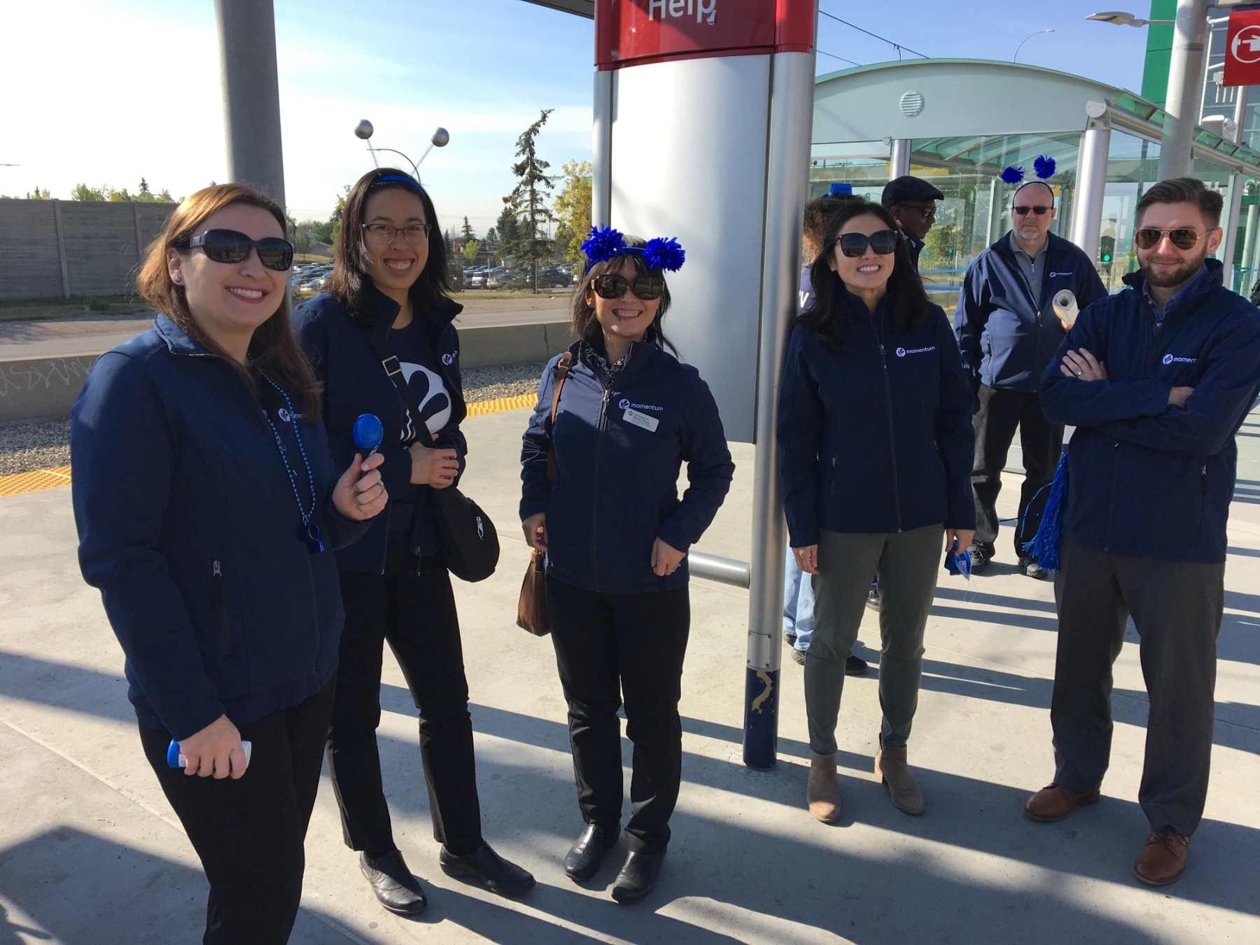 Six Momentum staff wearing blue jackets and pompom headbands stand together on a c-train platform.