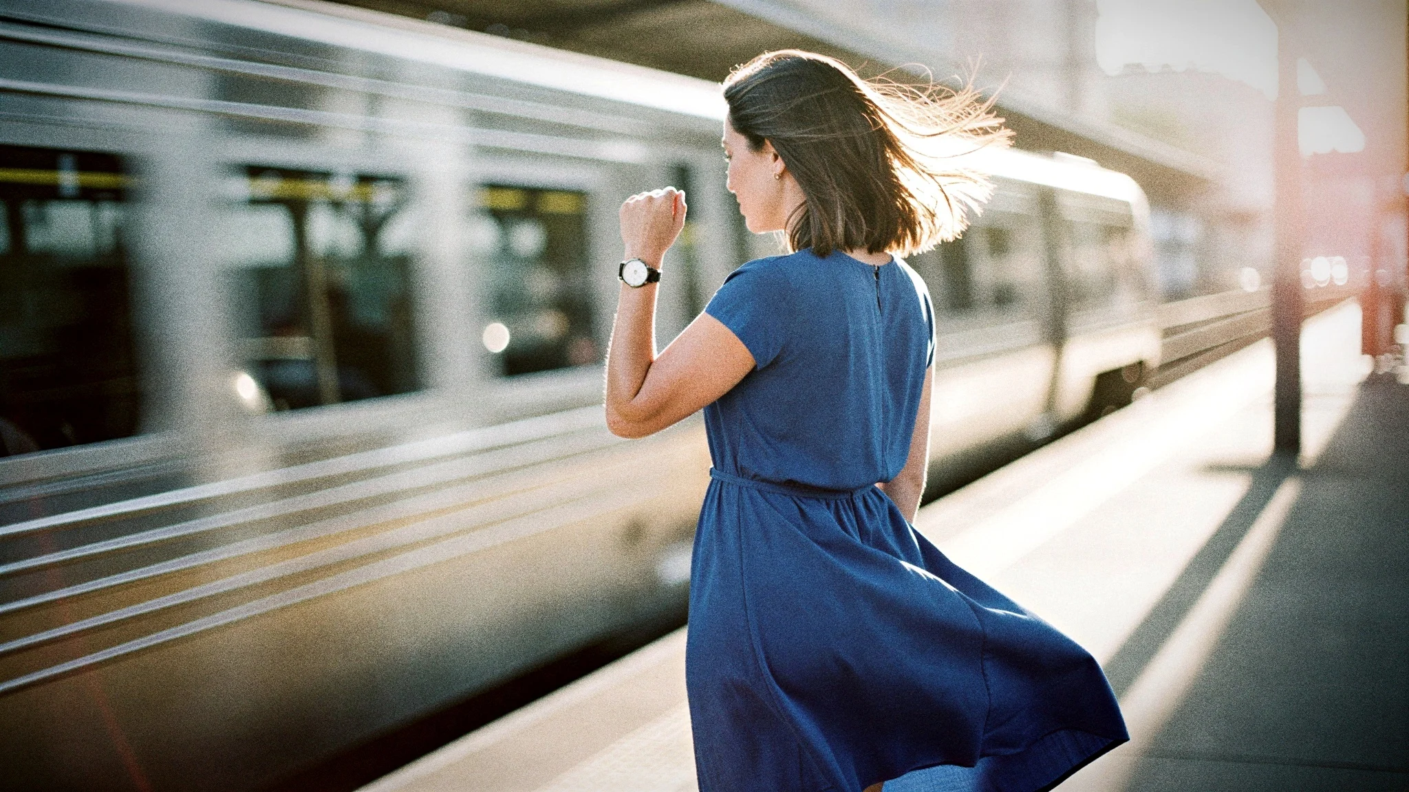 A woman in a blue dress checks her watch on a sunlit train platform as a train speeds past.