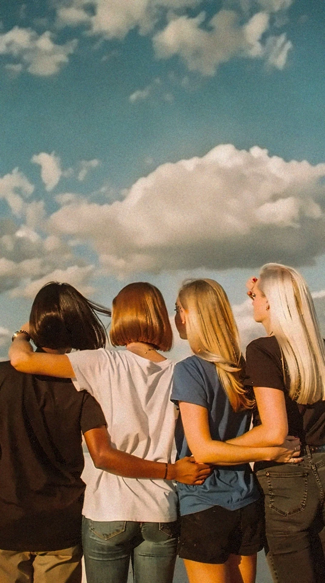Four women seen from behind on a rooftop, arms linked, looking out at a blue sky with clouds.