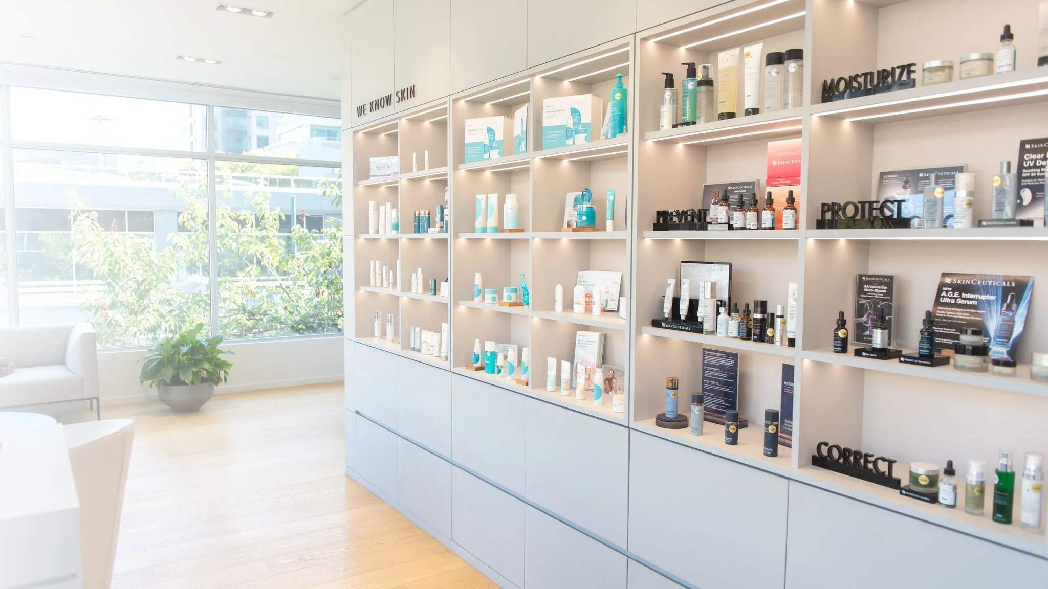 A lit clinic skincare shelving wall, its sections labelled Prevent, Protect, Correct and Moisturize, beside a full-height window.