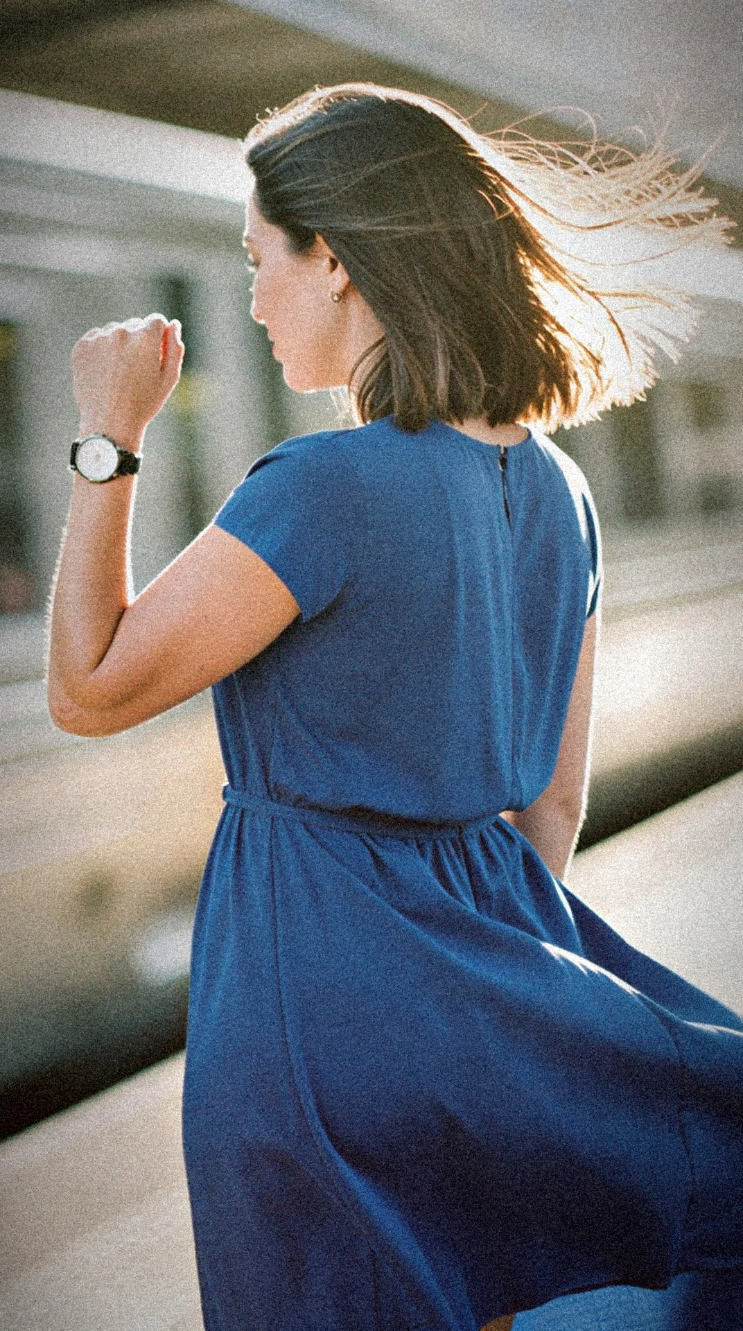 A woman in a blue dress checks her watch on a sunlit train platform, hair blowing in the wind.