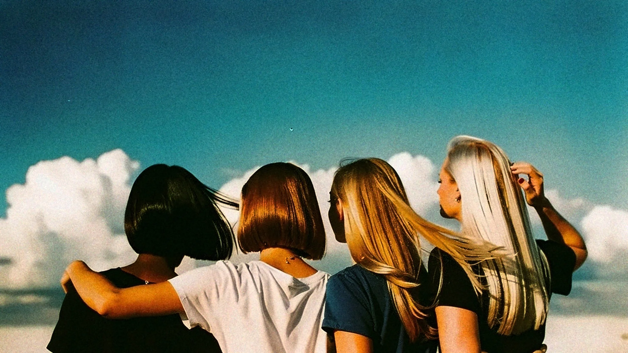Four women seen from behind on a city rooftop, arms around each other, hair blowing in the wind against a blue sky.